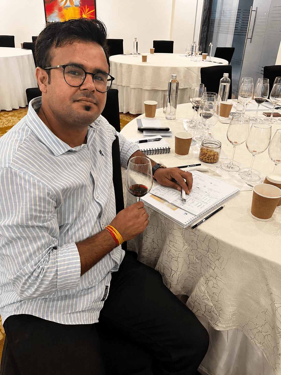 Man with glasses and striped shirt sitting at a table with wine glass, notebooks, and drinks in a banquet hall.