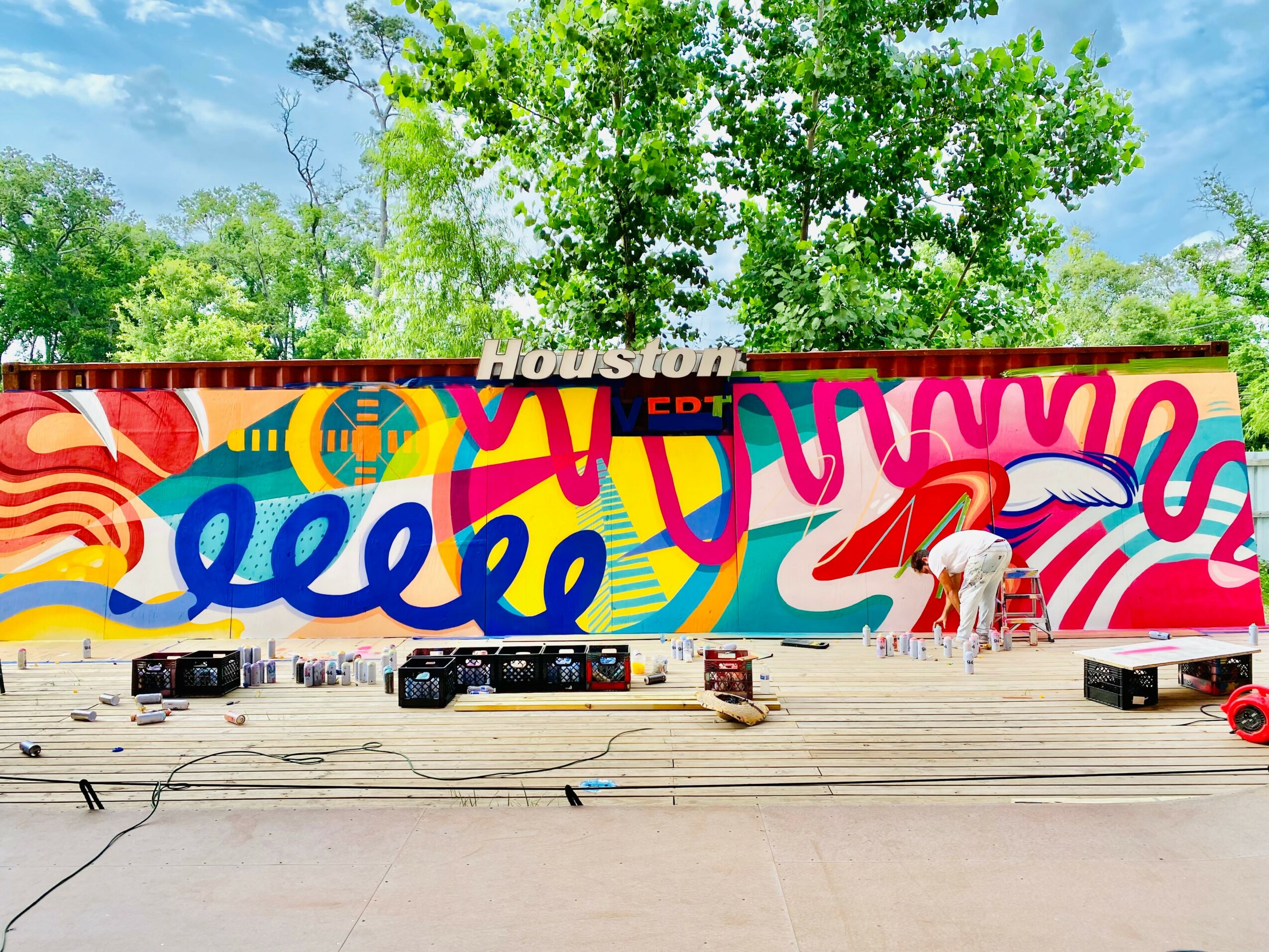 Colorful mural with abstract shapes and the word 'Houston' on a wall, with trees and sky in the background.
