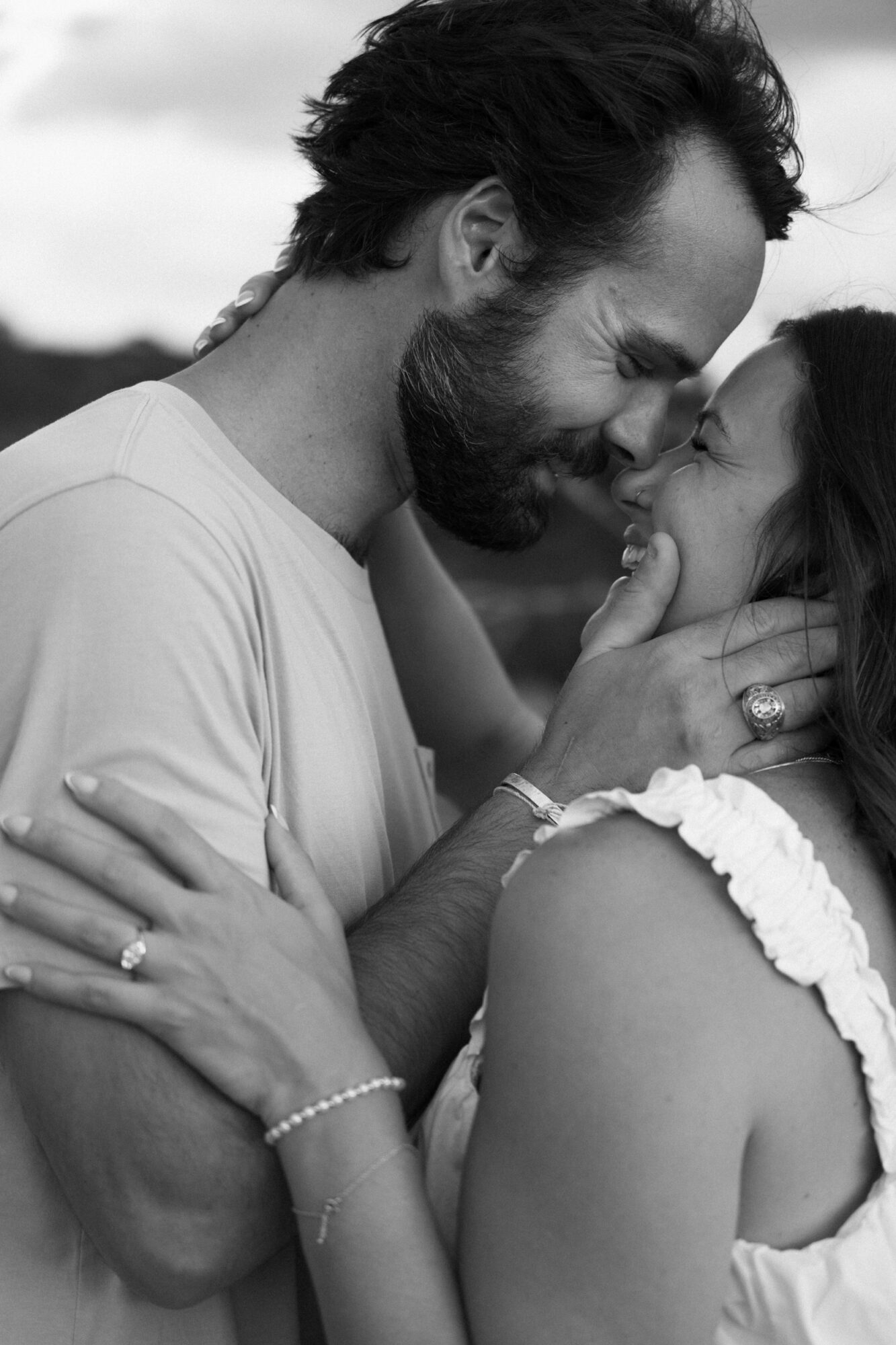 A man and woman face each other closely, smiling and touching faces, outdoors with a blurred background.