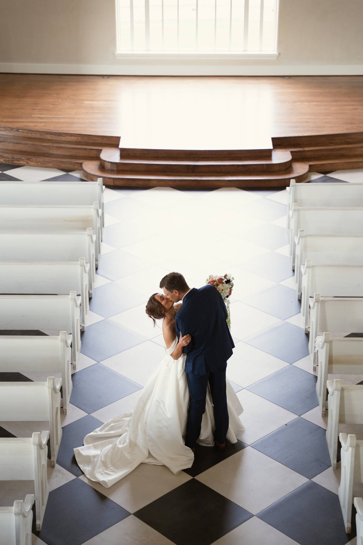 Bride and groom embrace in the aisle of a church with white pews and checkered floor, sunlight from window above.