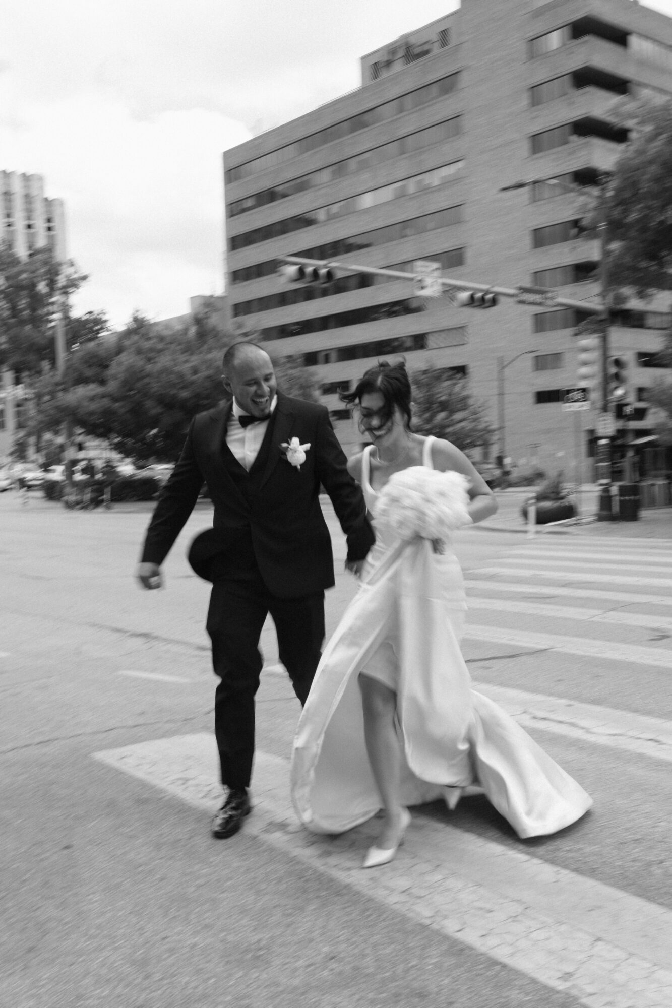 A couple in wedding attire walking together on a city street, holding hands and smiling.
