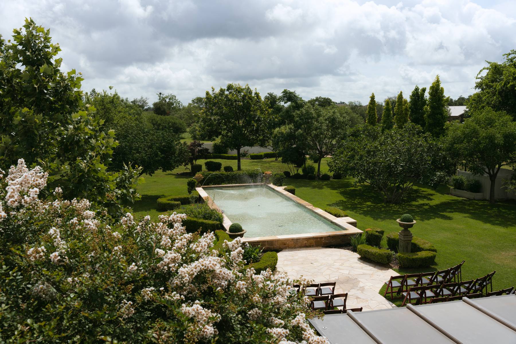 A landscaped garden with a rectangular water feature, surrounded by trees, benches, and cloudy sky.