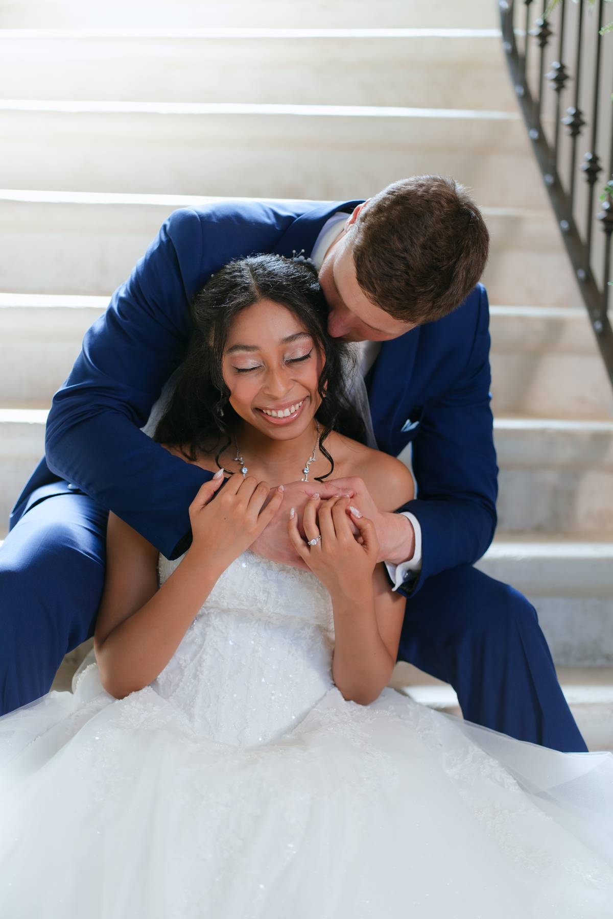 A smiling woman in a white dress sits on stairs, with a man in a blue suit embracing her from behind.