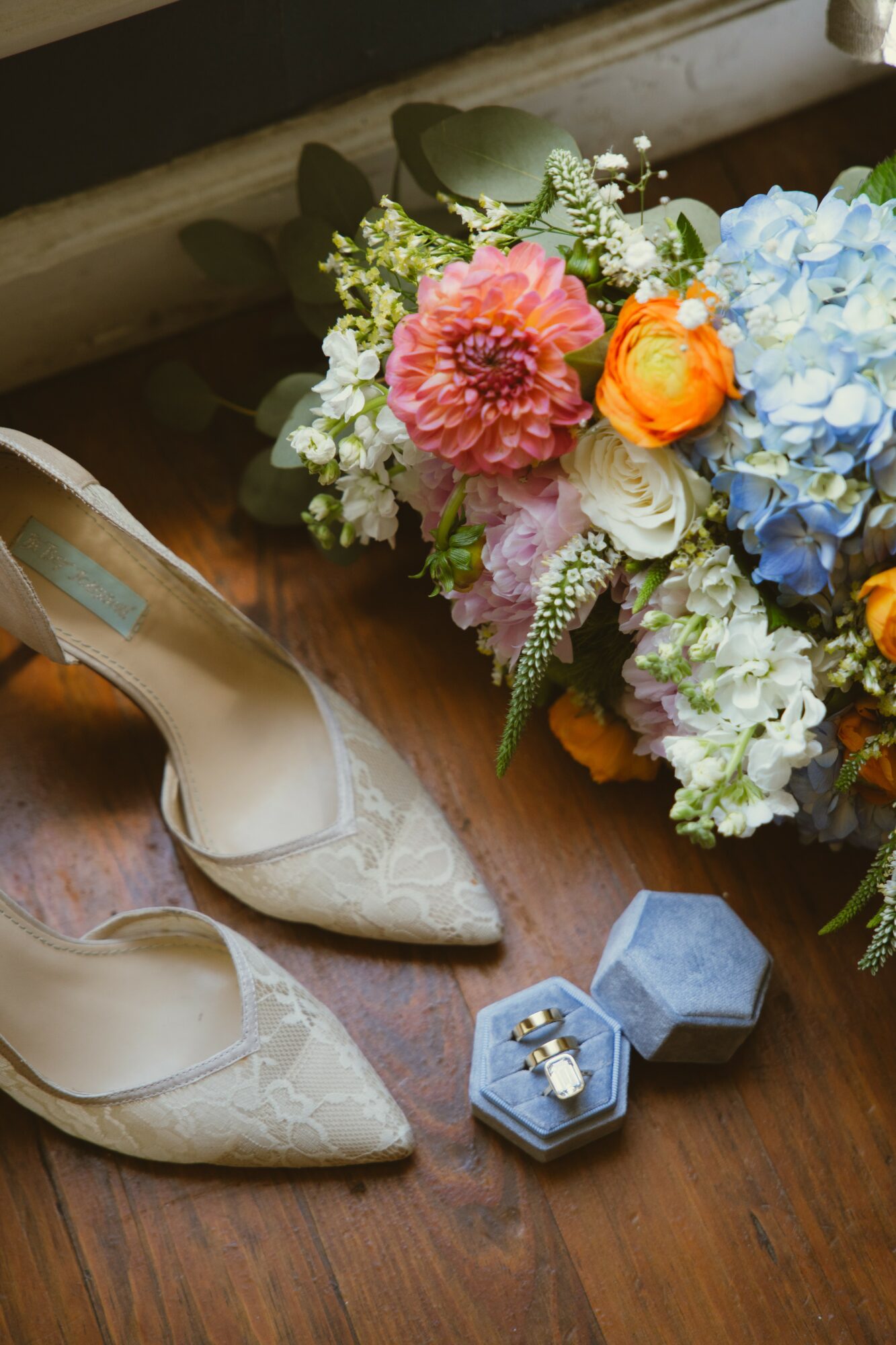 Pair of cream-colored high heels, a blue jewelry box with rings, and a colorful flower bouquet on a wooden surface.