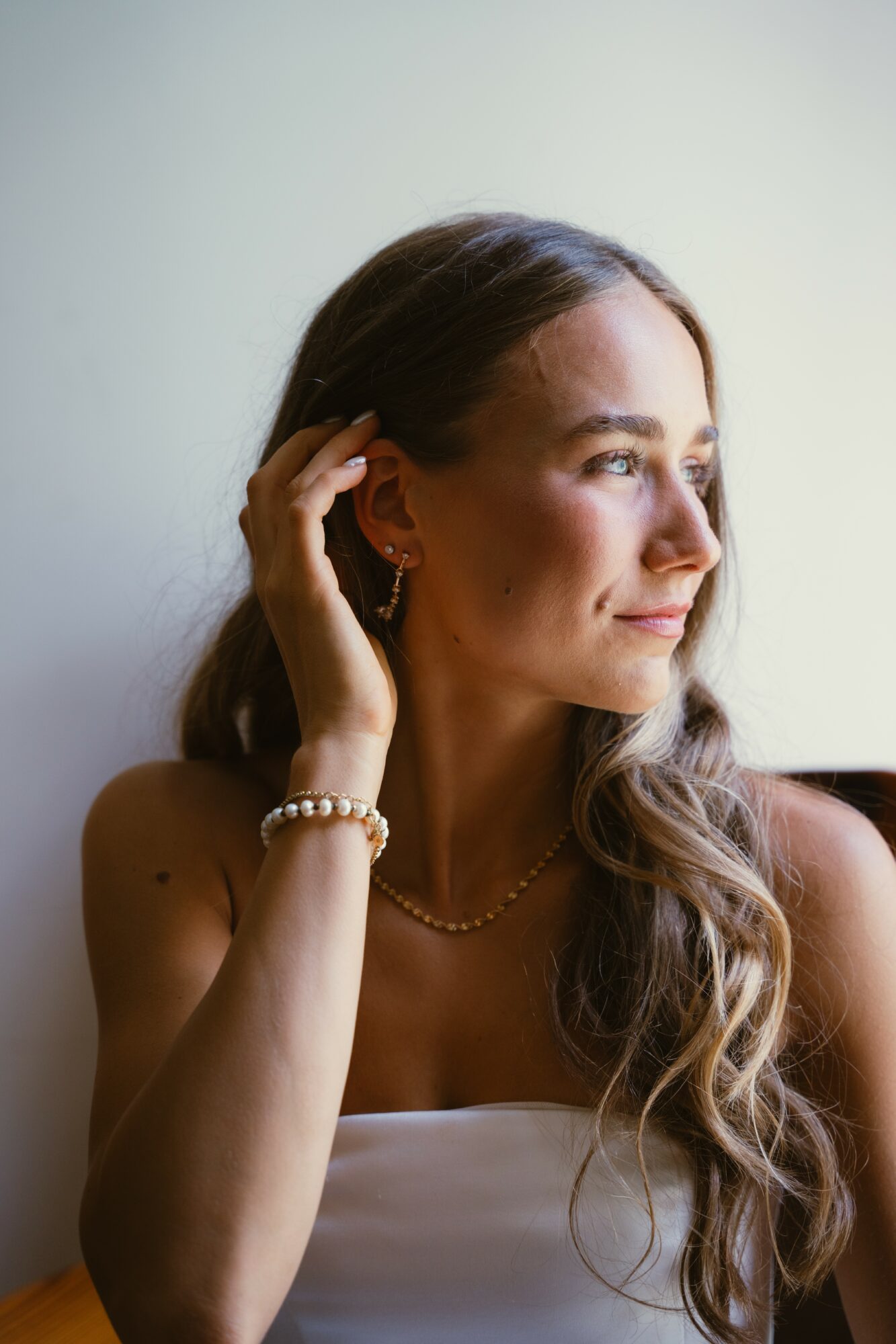 Woman with long wavy hair looking to the side, touching her ear, wearing jewelry, against a plain background.