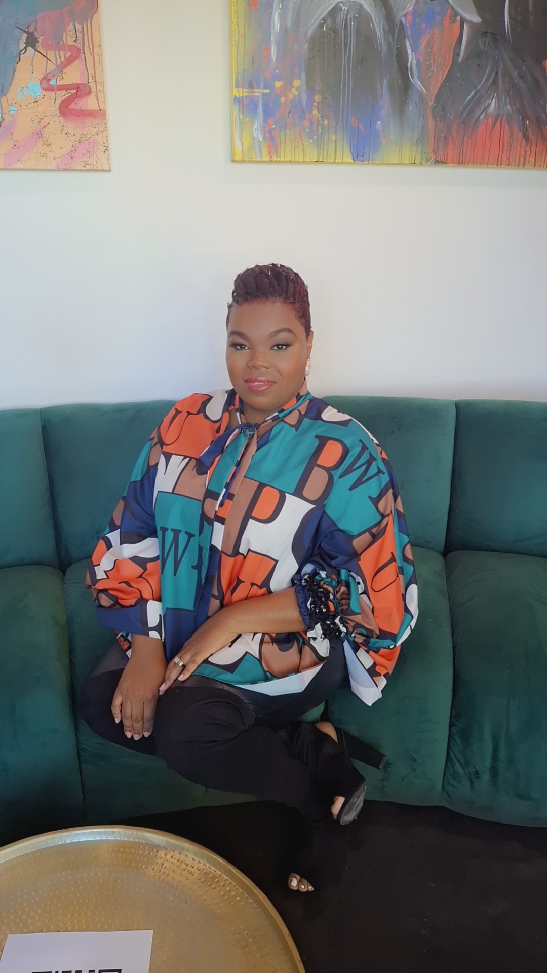 Woman sitting on a green sofa, wearing a colorful patterned blouse, with abstract paintings on the wall behind her.