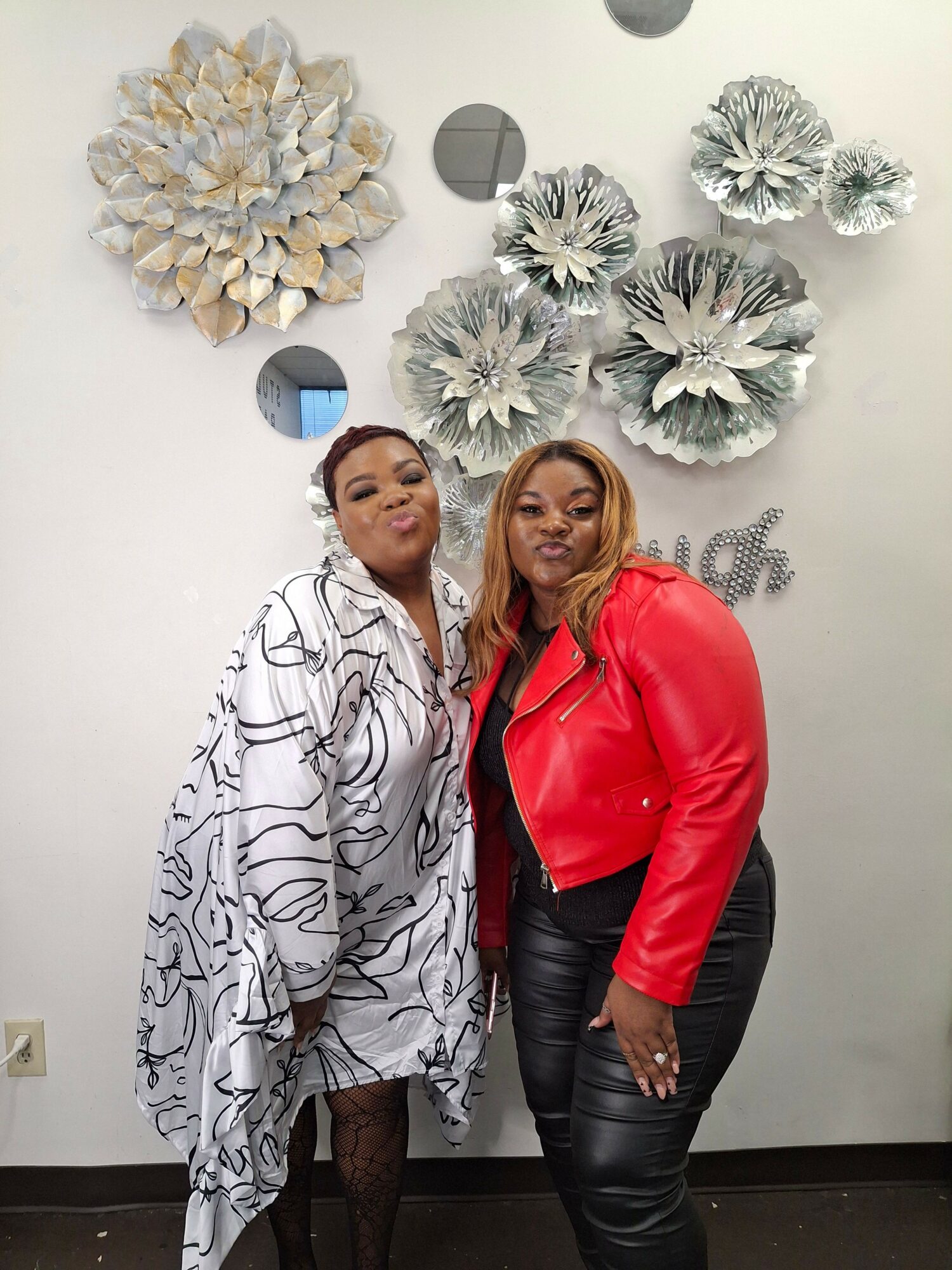 Two women stand in front of a white wall decorated with large silver and white floral art pieces, posing for a photo.