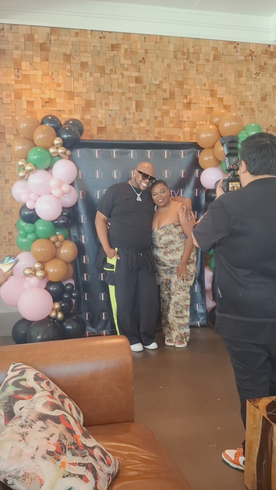 Two people pose in front of a decorated backdrop with balloons, with a person taking their photo, in an indoor setting.