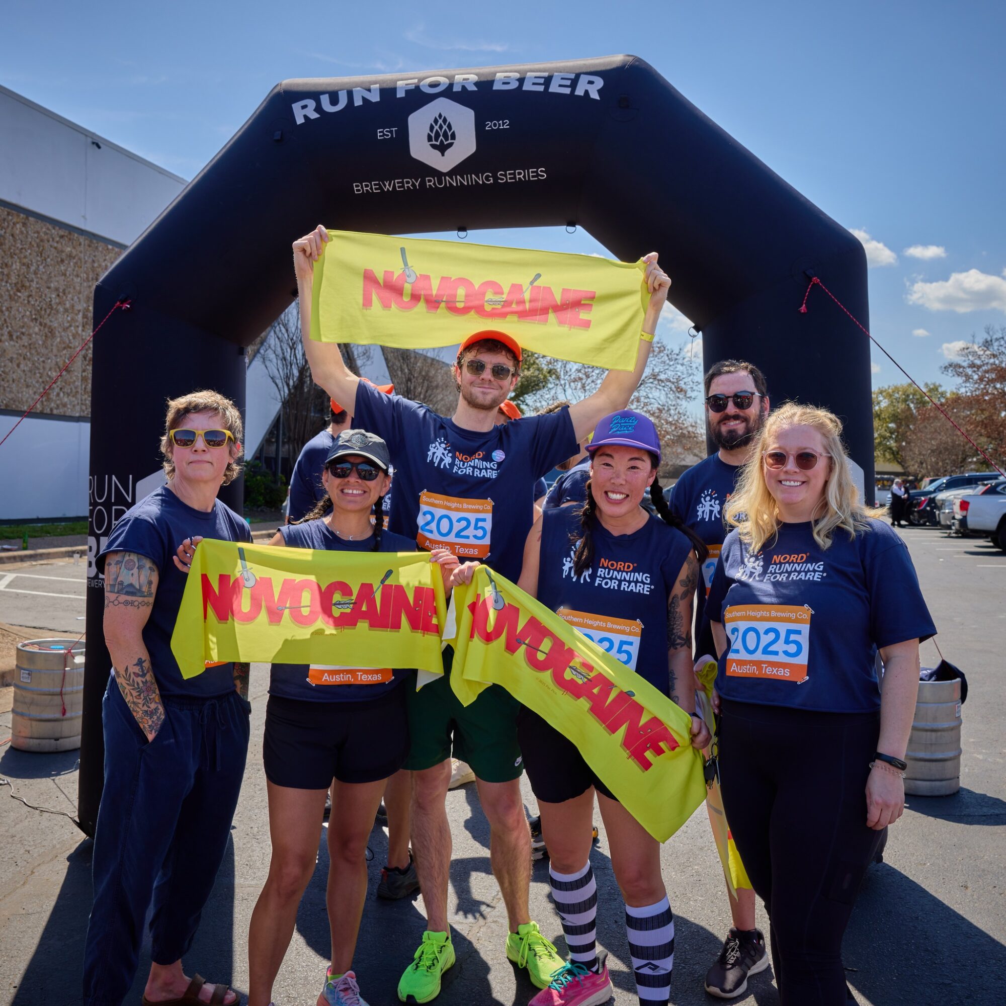 Group of six people holding yellow banners with red text, standing under a black inflatable arch at a running event, sunny day.