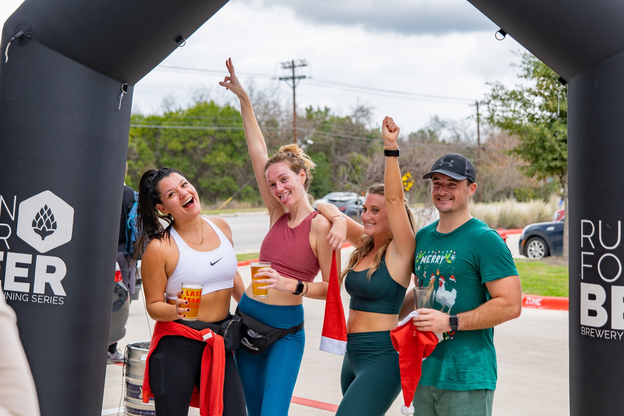 Four people celebrating outdoors under an inflatable arch, holding drinks, smiling, and raising their hands.