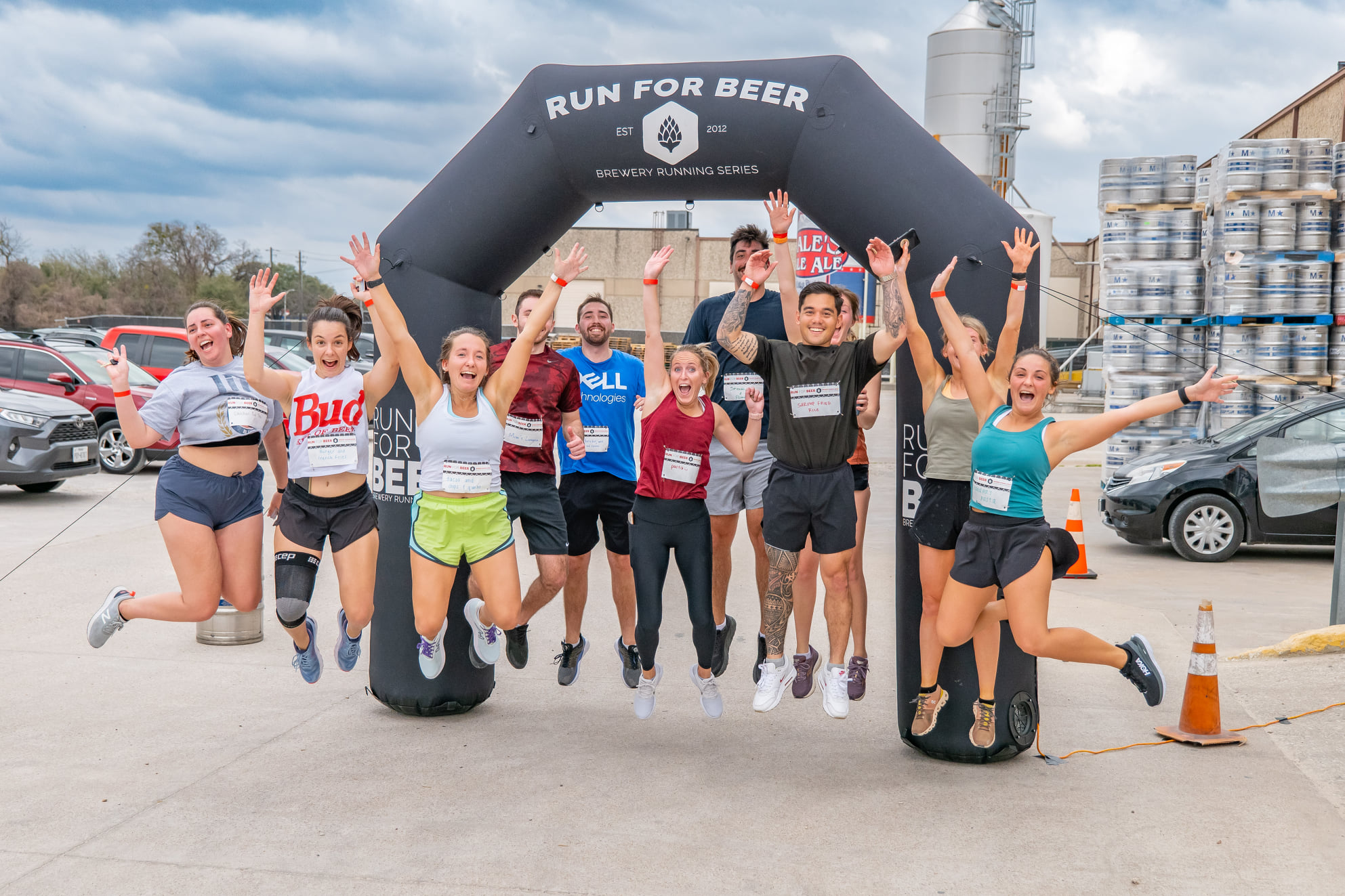 Group of people jumping in front of an inflatable arch that says 'Run for Beer' outdoors, with parked cars and buildings in background.