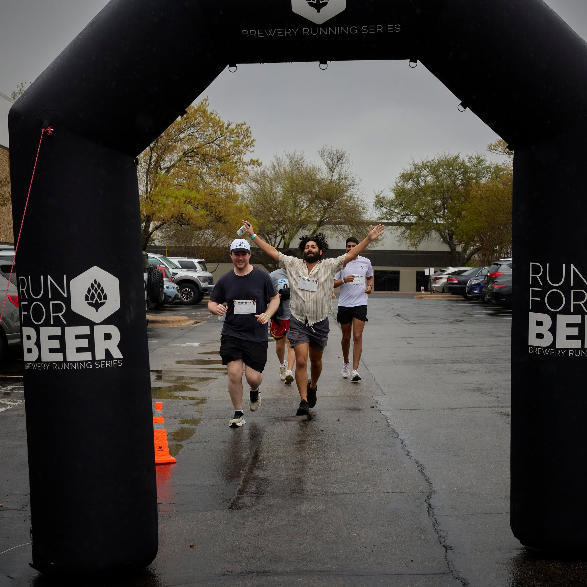 People running through a finish line arch in a parking lot, with trees and cars in the background.