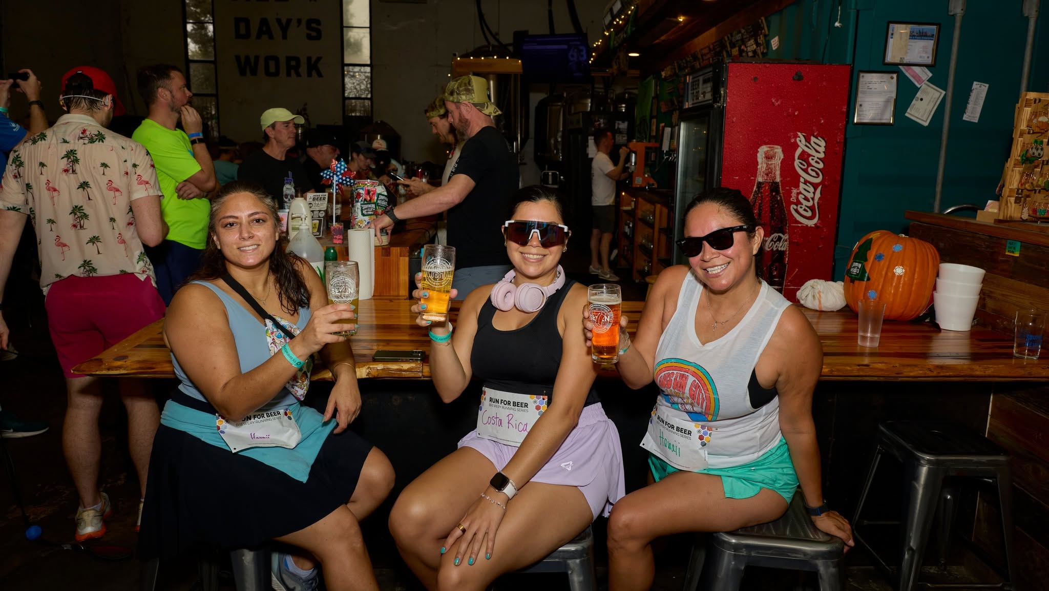 Three women sitting at a bar holding drinks, with people and decorations in the background.