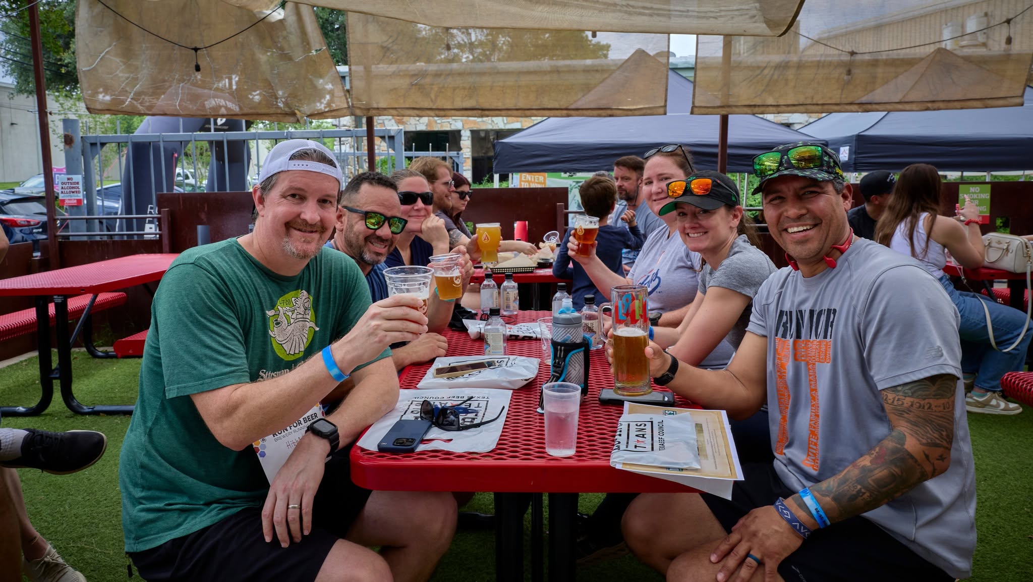 Group of people sitting at a table outdoors, smiling, holding drinks, with tents and trees in the background.