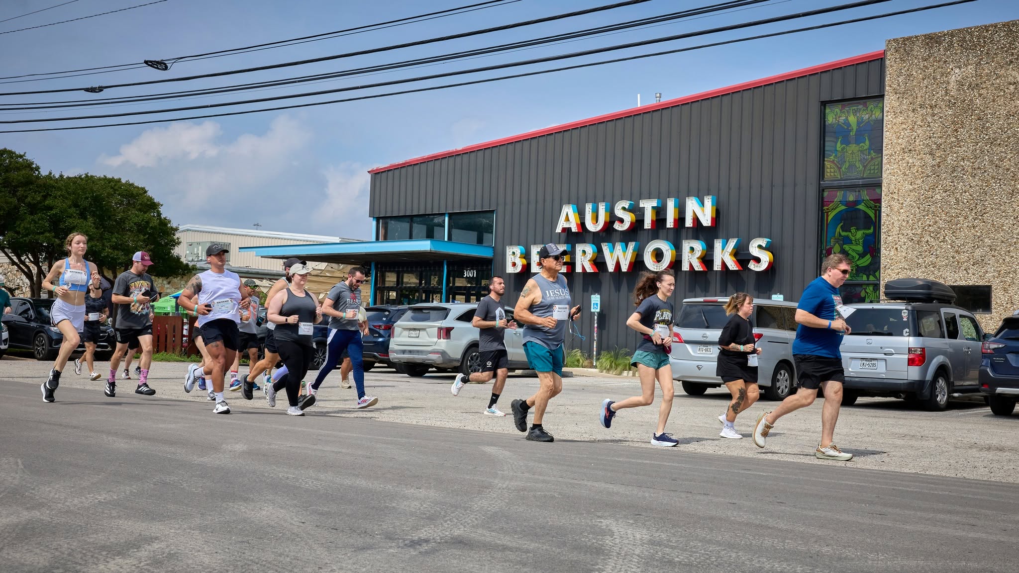 Group of people running outside a building with a sign reading 'AUSTIN SKYWORKS'. Cars parked nearby, clear sky.
