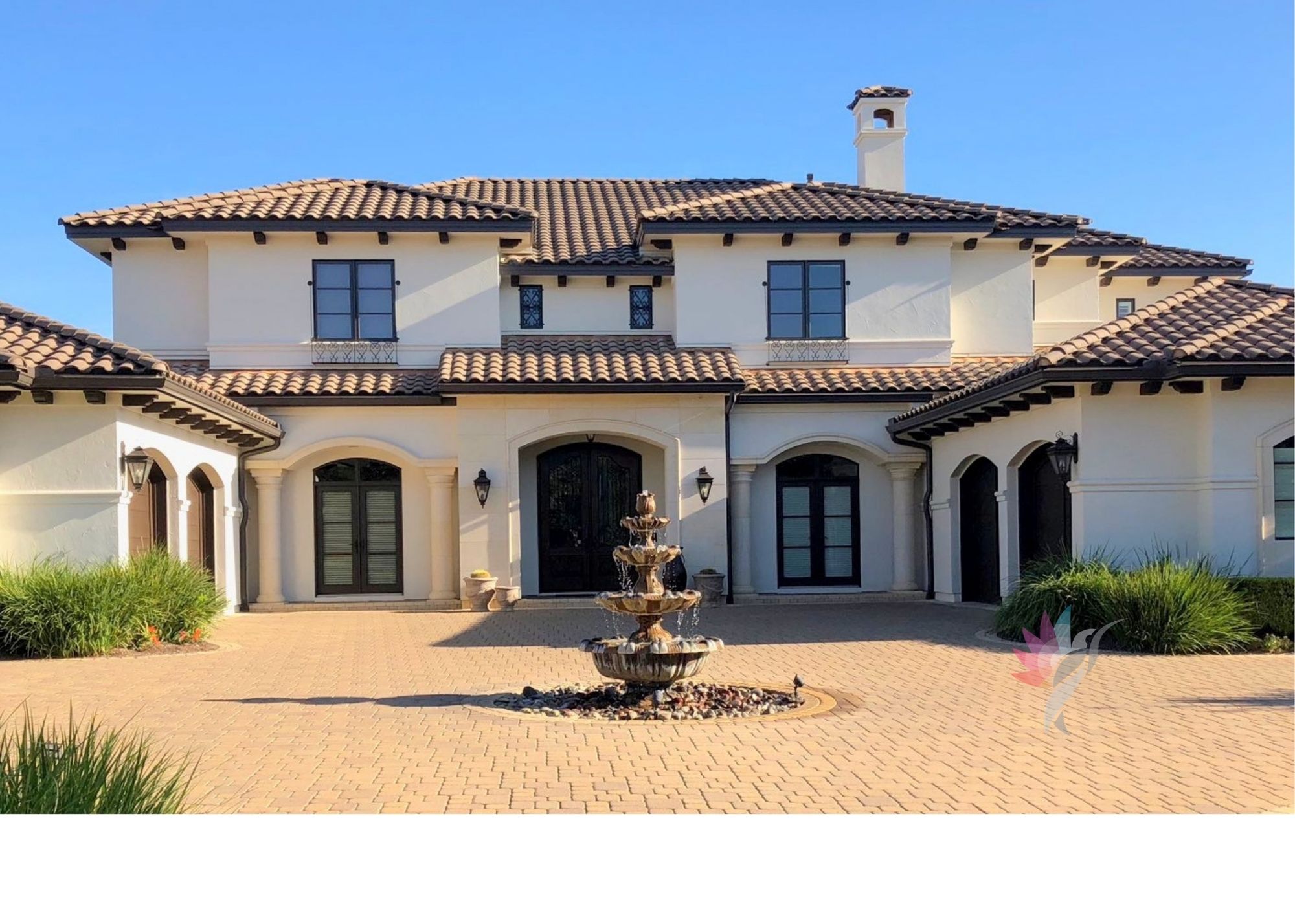 Large white house with a fountain in front, multiple windows, and a tiled roof, under a clear blue sky.