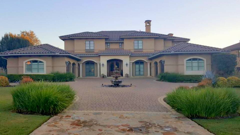 Large house with a driveway, fountain, and landscaped bushes in front, under a clear sky.