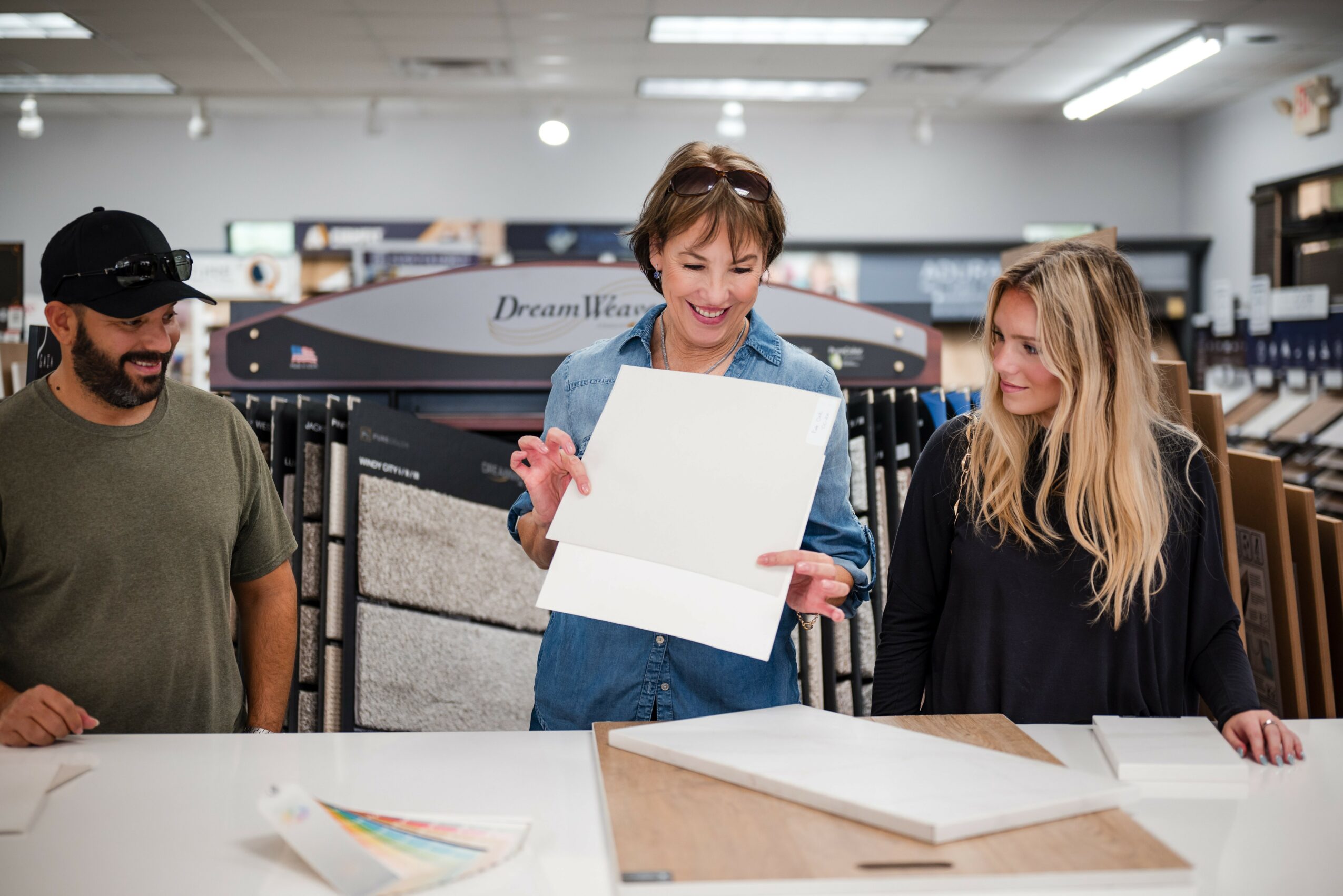 Three people in a store, woman showing a white box to two others, with flooring samples and display racks in background.