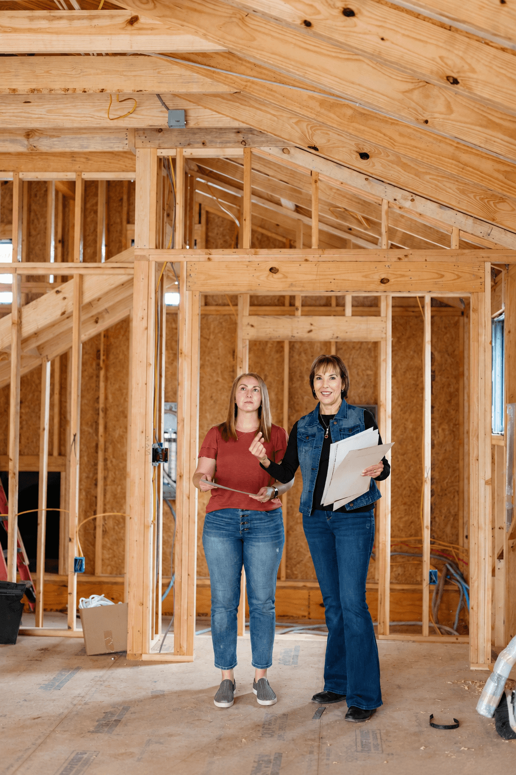 Two women stand inside a wooden house under construction, holding blueprints and smiling, with unfinished walls and ceiling visible.