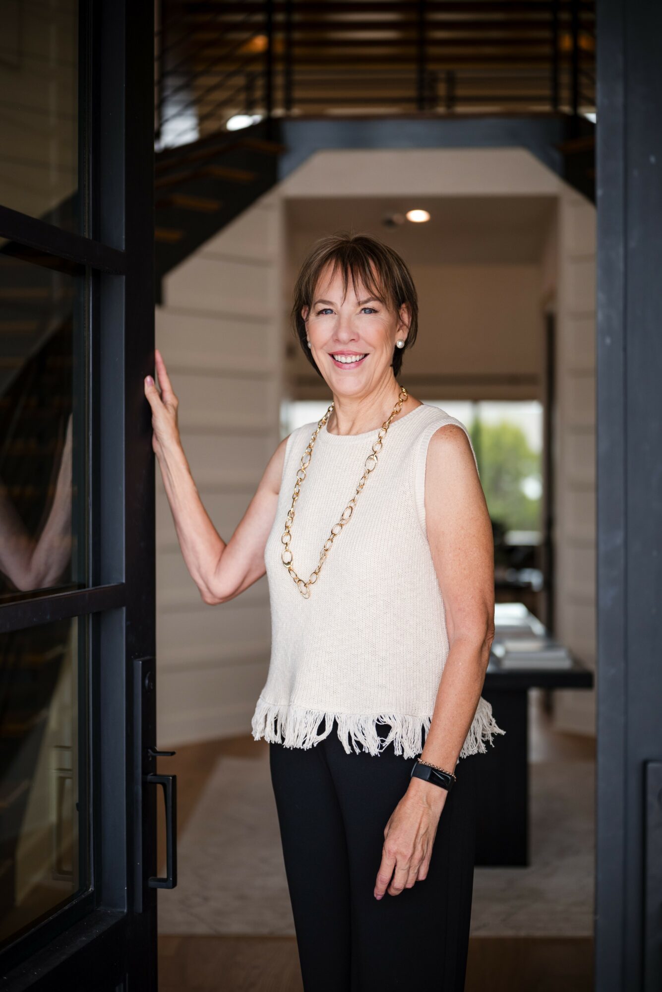 Woman standing in doorway, smiling, wearing a white sleeveless top with fringe, black pants, and a necklace.