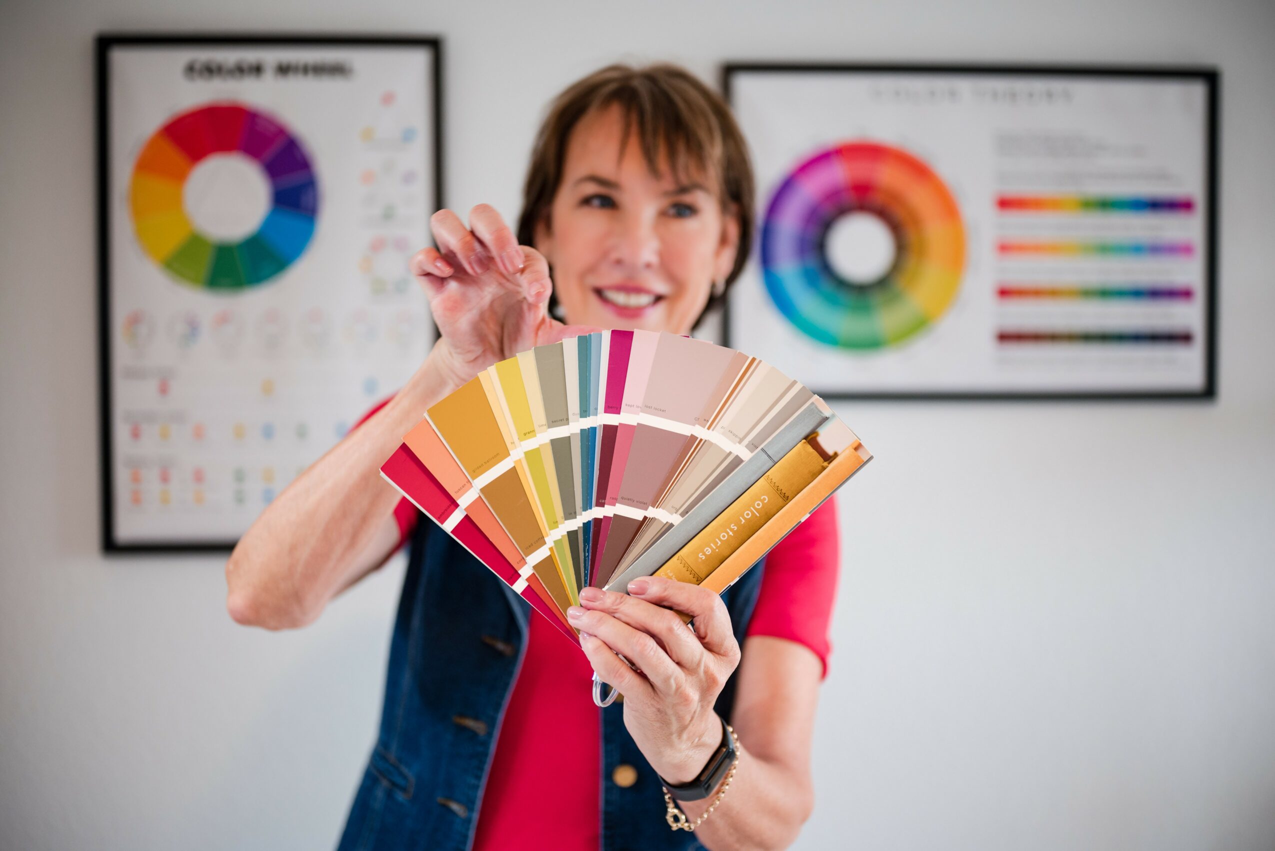 Woman holding color swatches in front of color charts on wall, smiling, in a bright room.