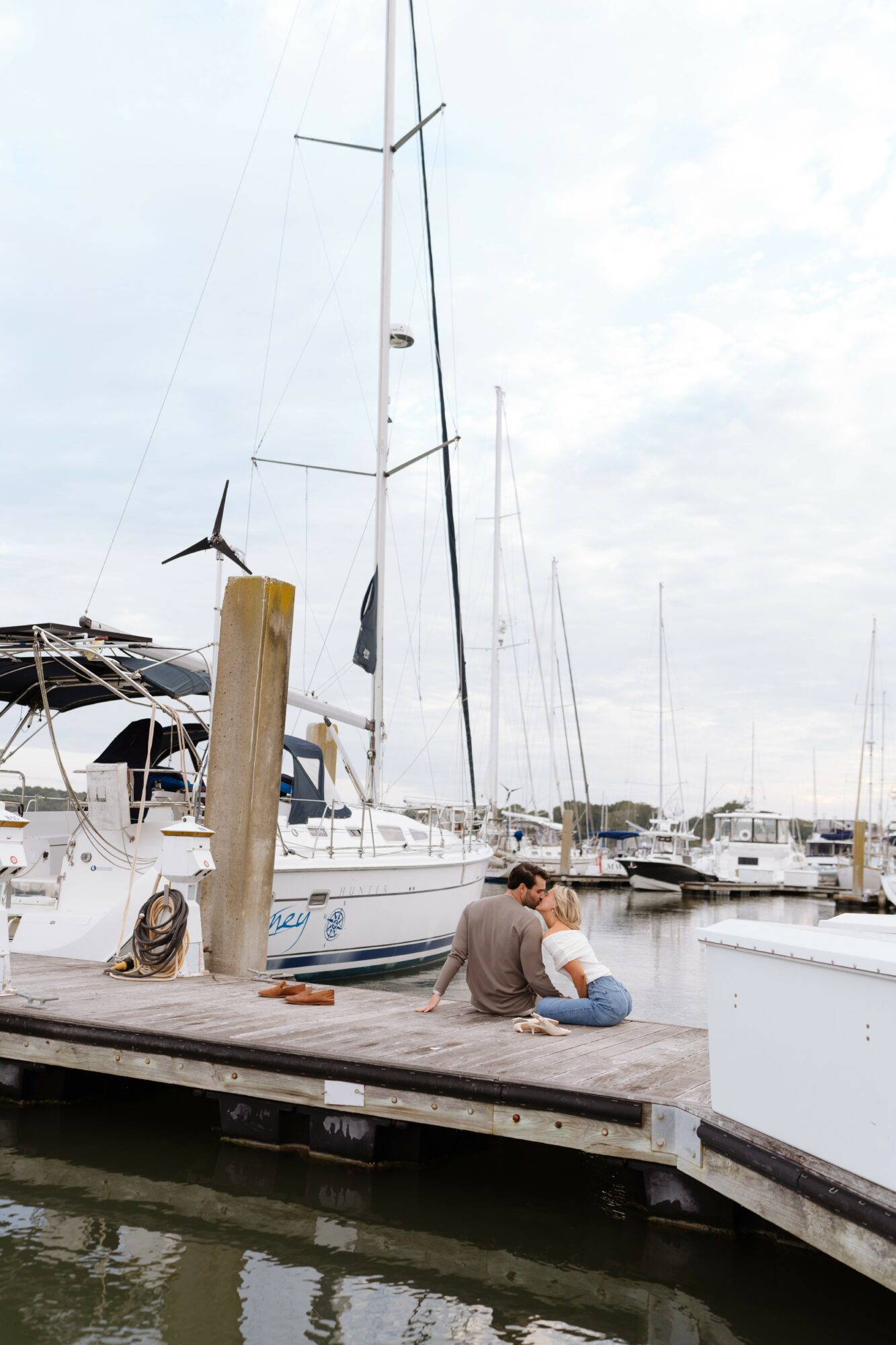 Two people sit on a dock near a sailboat at a marina, with other boats and a cloudy sky in the background.