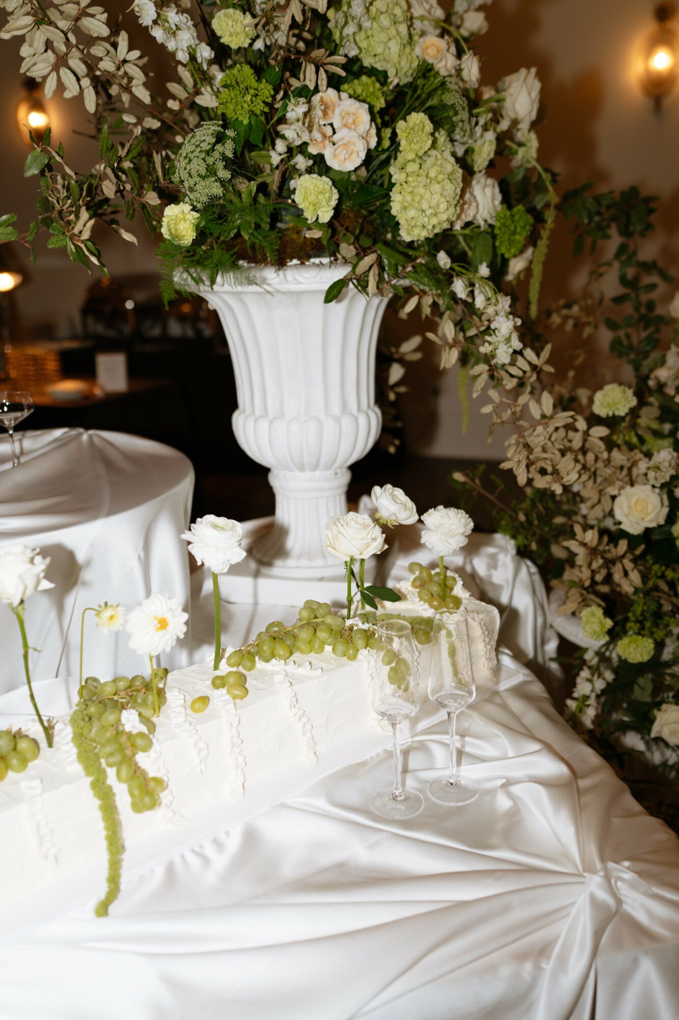 Elegant floral arrangement with white and green flowers in a tall white vase on a satin tablecloth, with additional flowers and candles in background.