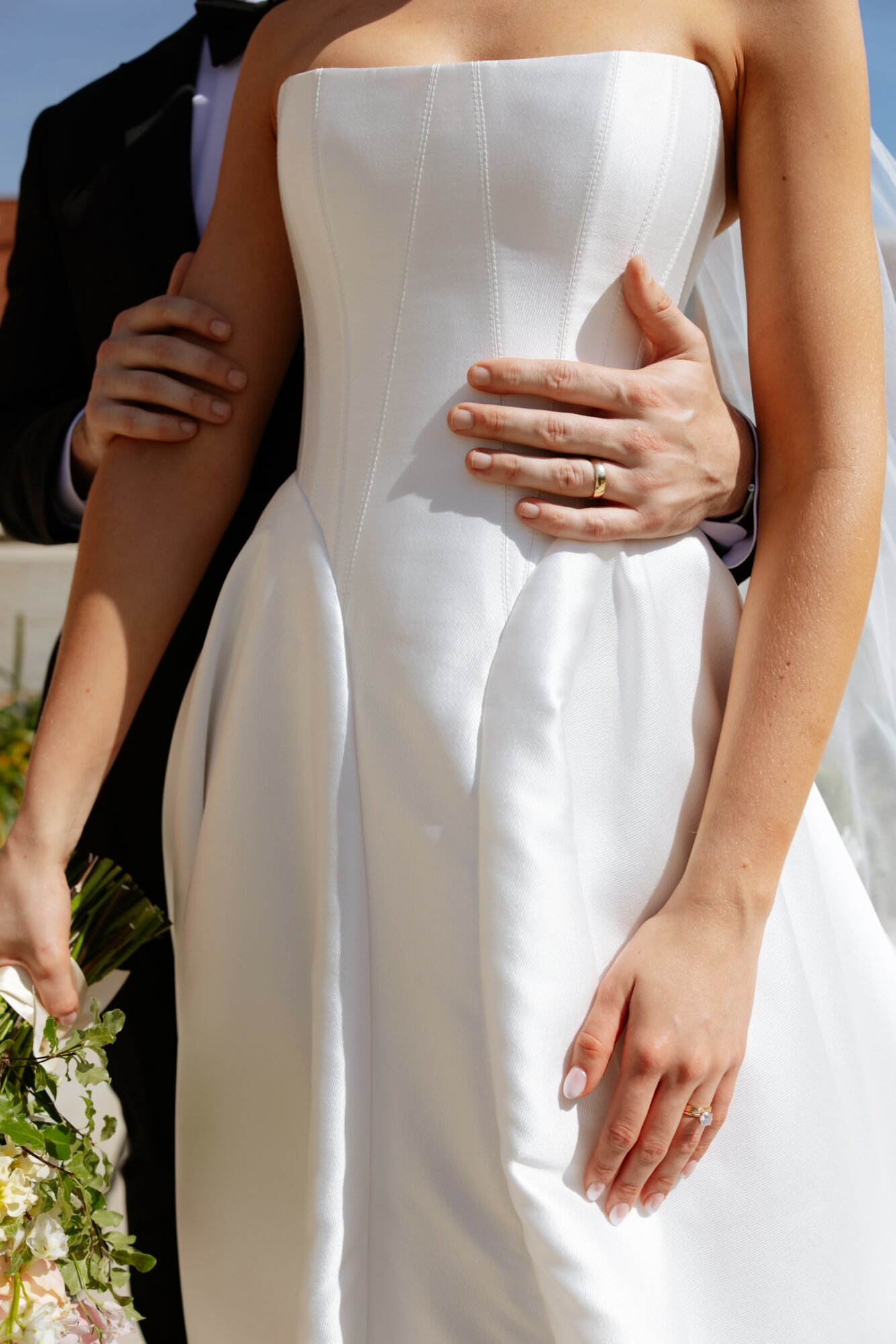 Close-up of a bride in a strapless white wedding dress with a groom's hand on her waist, holding a bouquet.