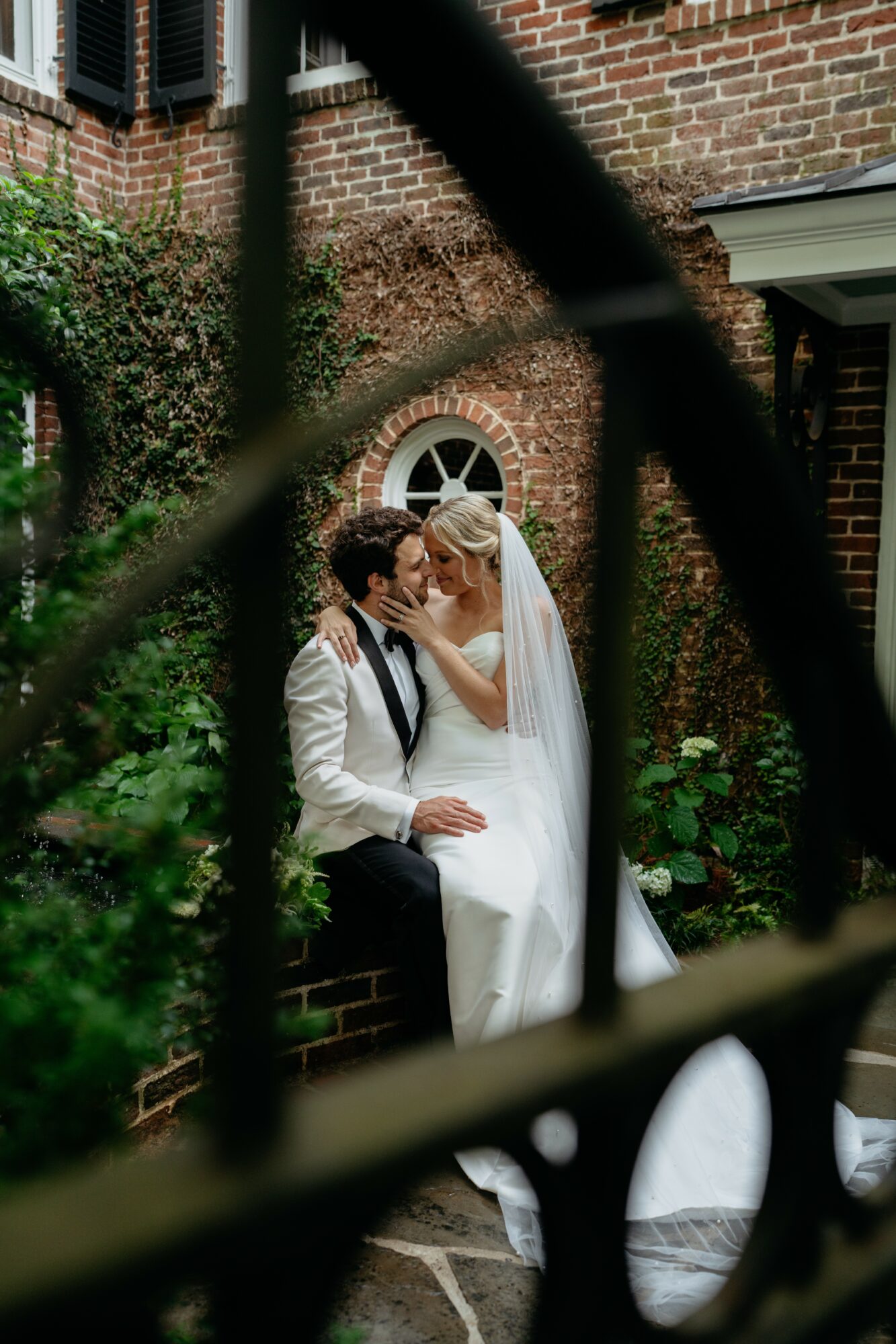 Bride and groom sitting close, sharing a kiss outdoors near a brick building with a small arched window, surrounded by greenery.