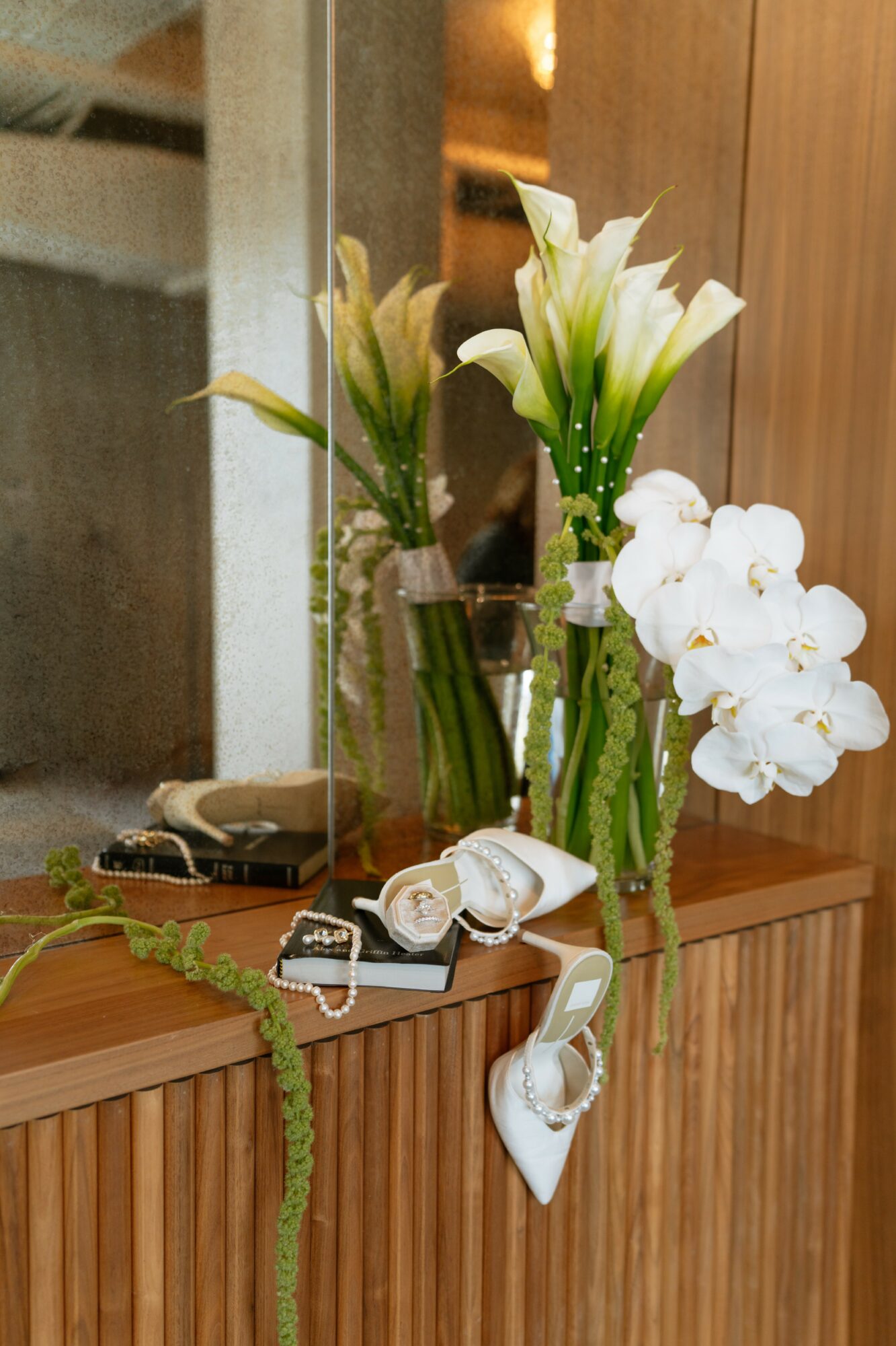 Three vases with white lilies and orchids on a wooden surface, with watches and a heart-shaped ornament hanging below.