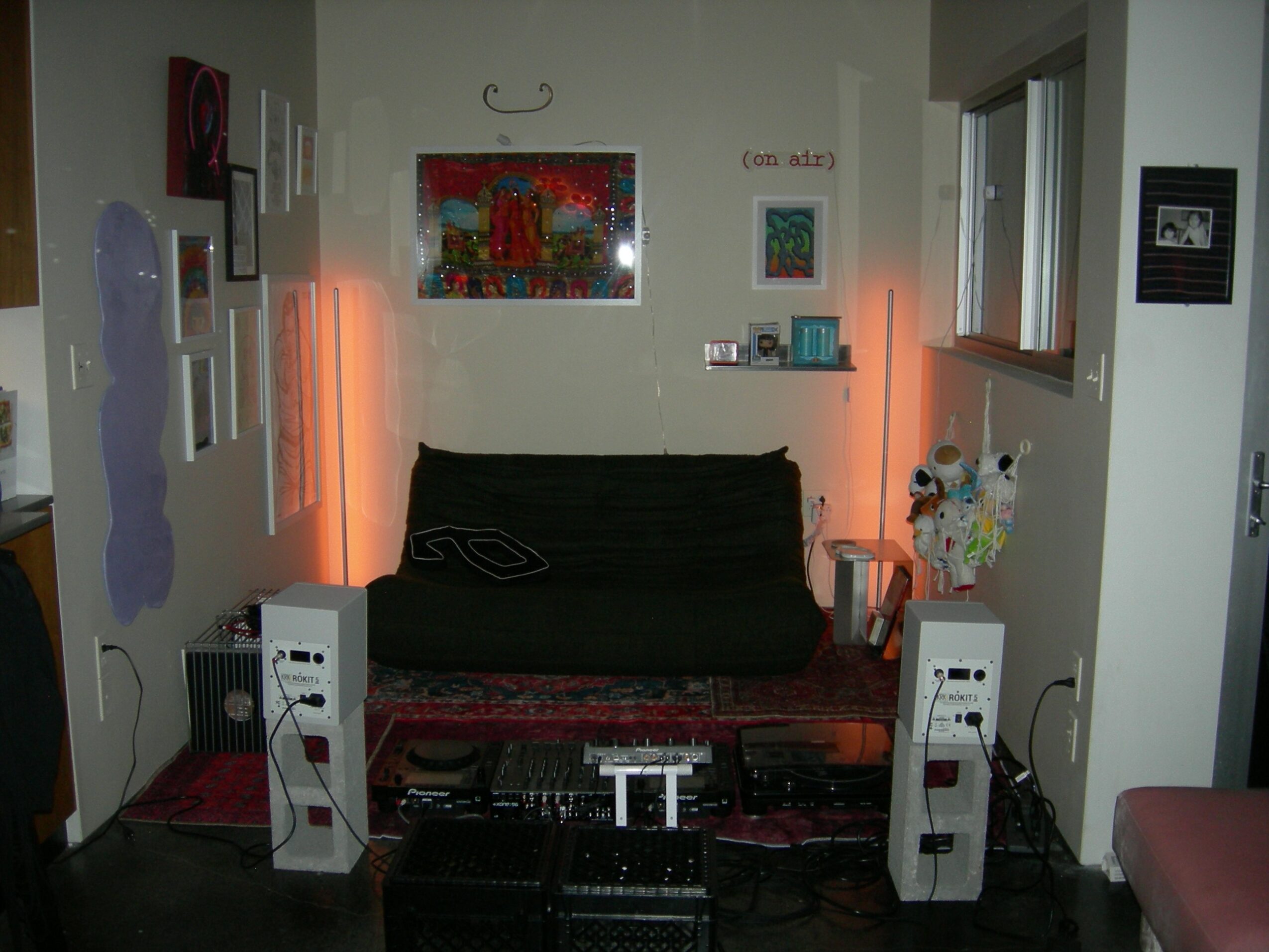 Living room with black sofa, colorful artwork, and two white speakers, illuminated by orange lights, with a window on the right.