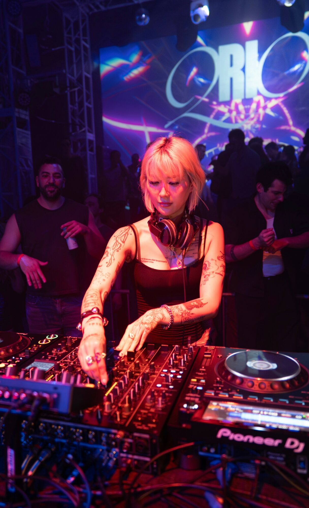 Female DJ with blonde hair and headphones performing at a nightclub with a crowd and colorful lights.