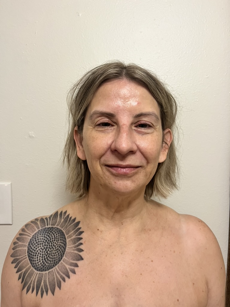 Woman with short, light brown hair and a sunflower tattoo on her shoulder, standing against a plain wall.