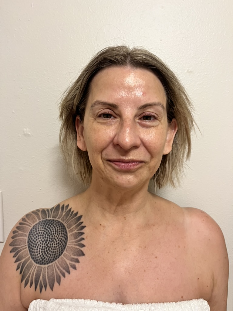 Woman with short hair and a sunflower tattoo on her shoulder, standing against a plain wall.