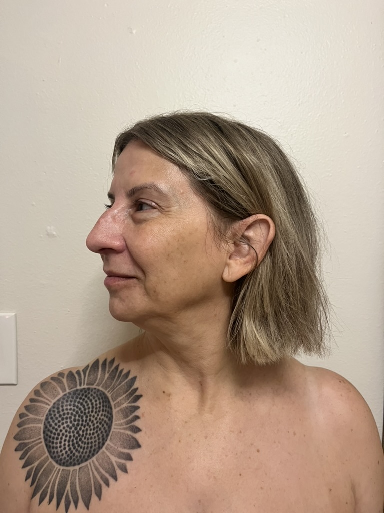 Side profile of a woman with shoulder-length blonde hair and a sunflower tattoo on her shoulder, against a plain wall.