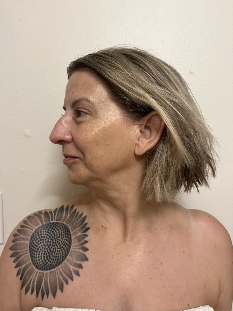 Side profile of a woman with short, layered hair and a sunflower tattoo on her shoulder, against a plain wall.