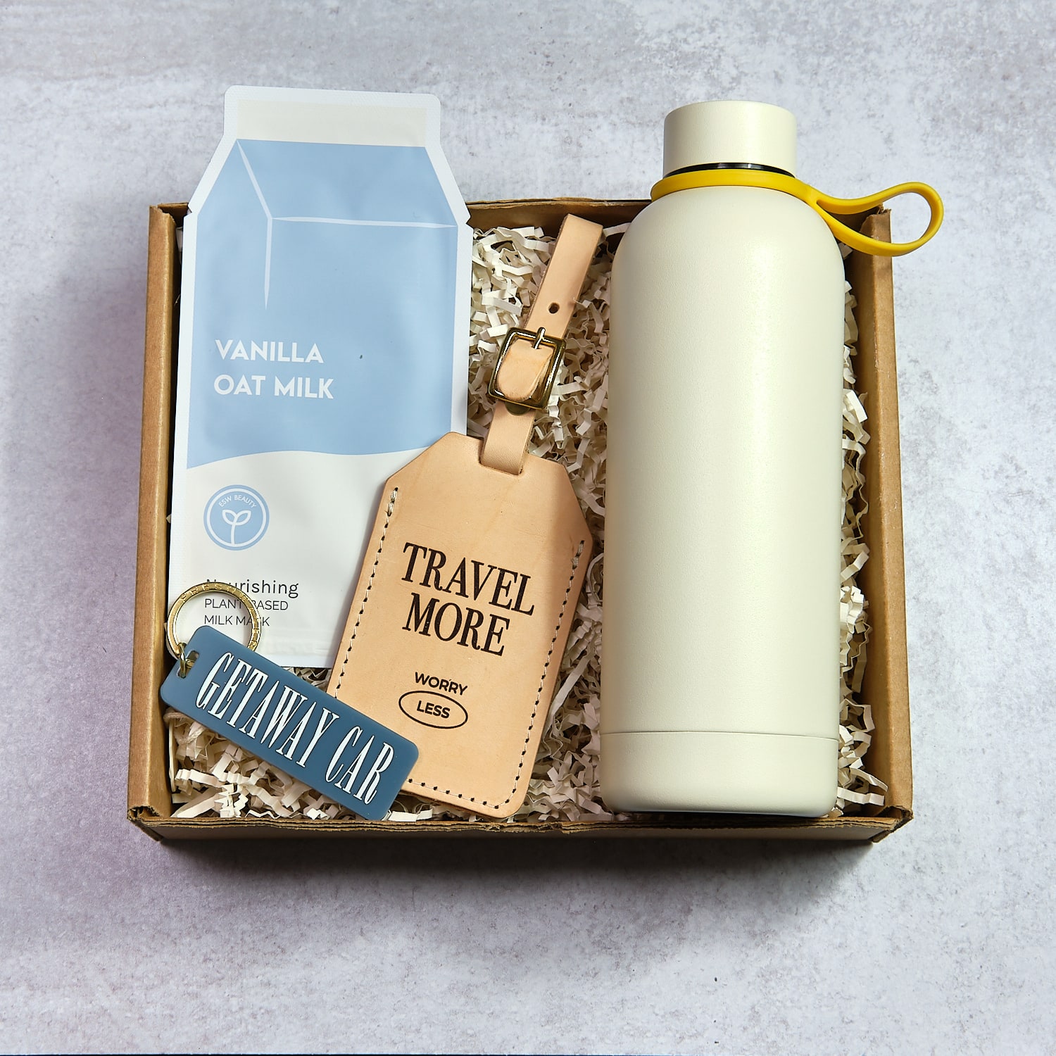 Open box with vanilla oat milk, travel tag, water bottle, and keychain on shredded paper, on gray surface.
