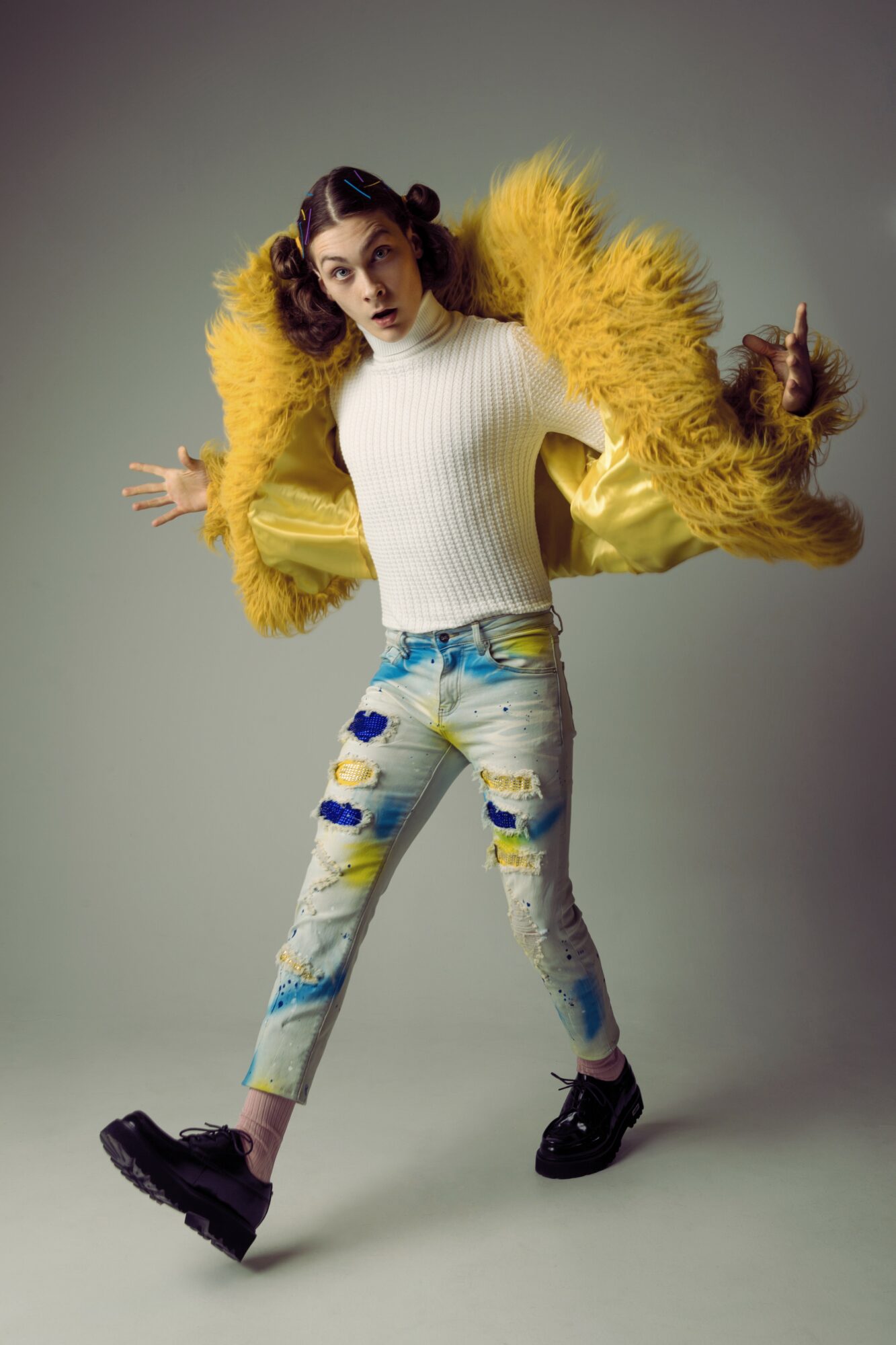 Person wearing a yellow furry coat, white sweater, and ripped jeans, standing with arms outstretched, against a plain background.