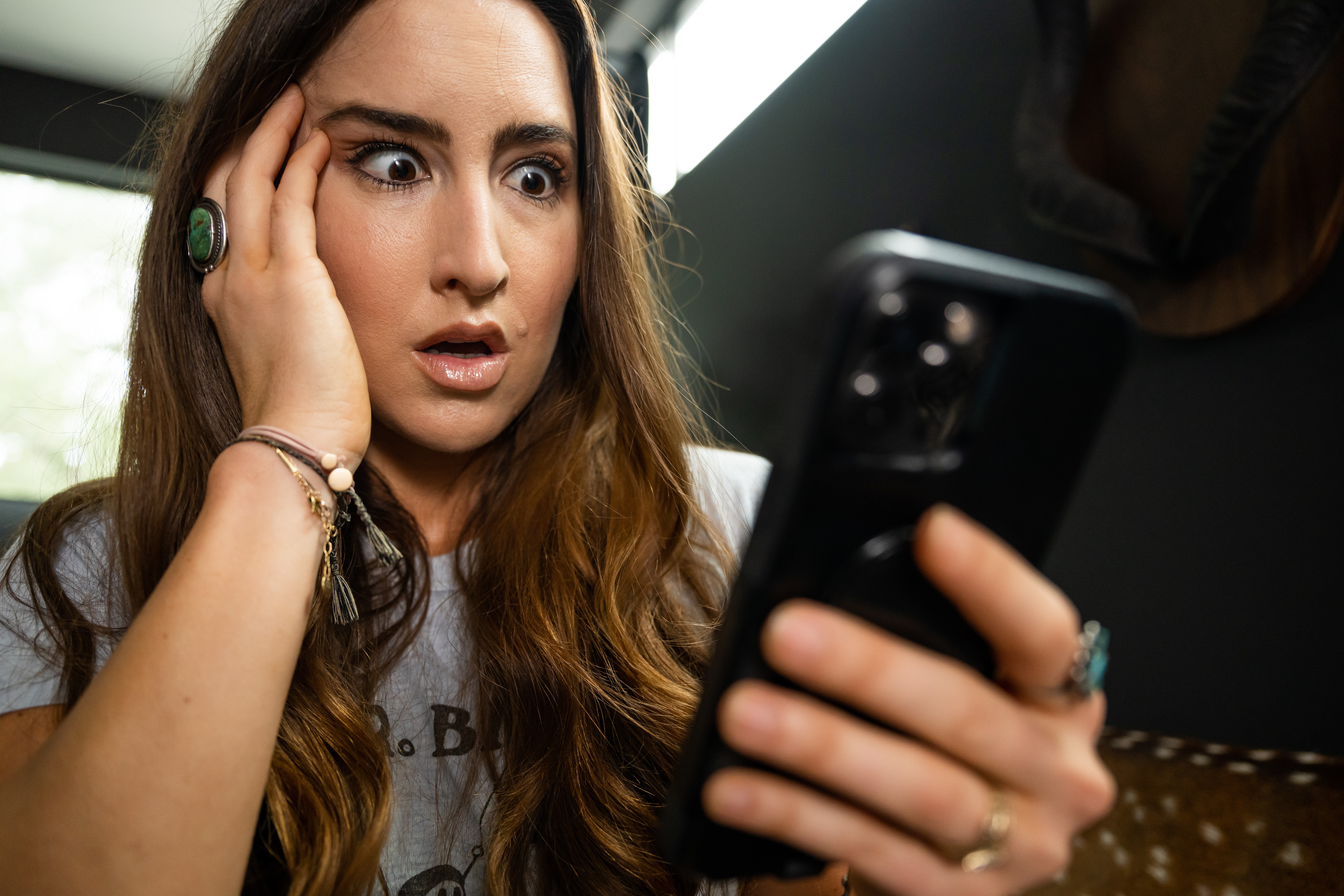 Young woman with long hair looks surprised at her phone, hand on her face, indoors with natural light.