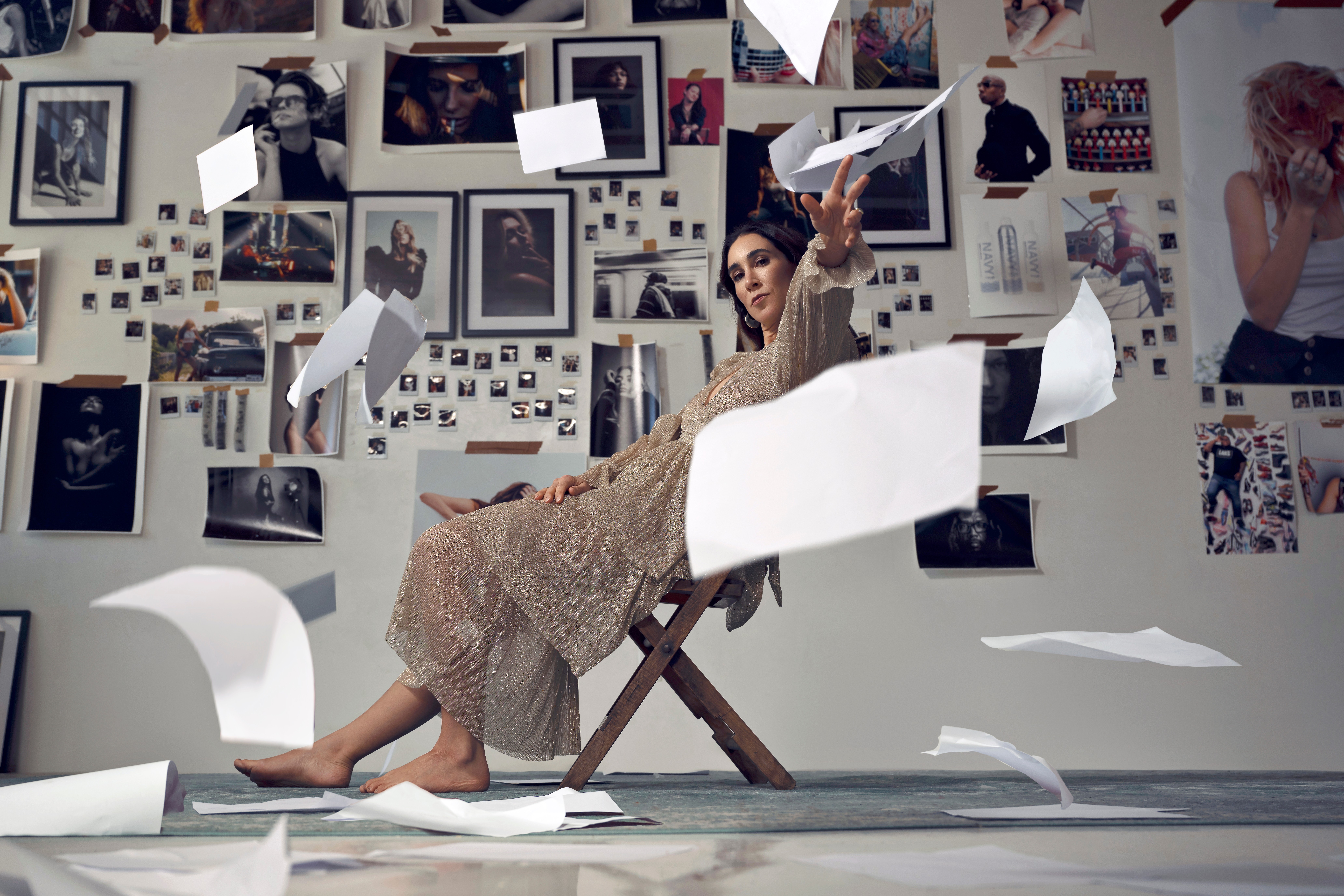 Woman sitting on a chair in a room with walls covered in photos, papers flying around her, some on the floor.