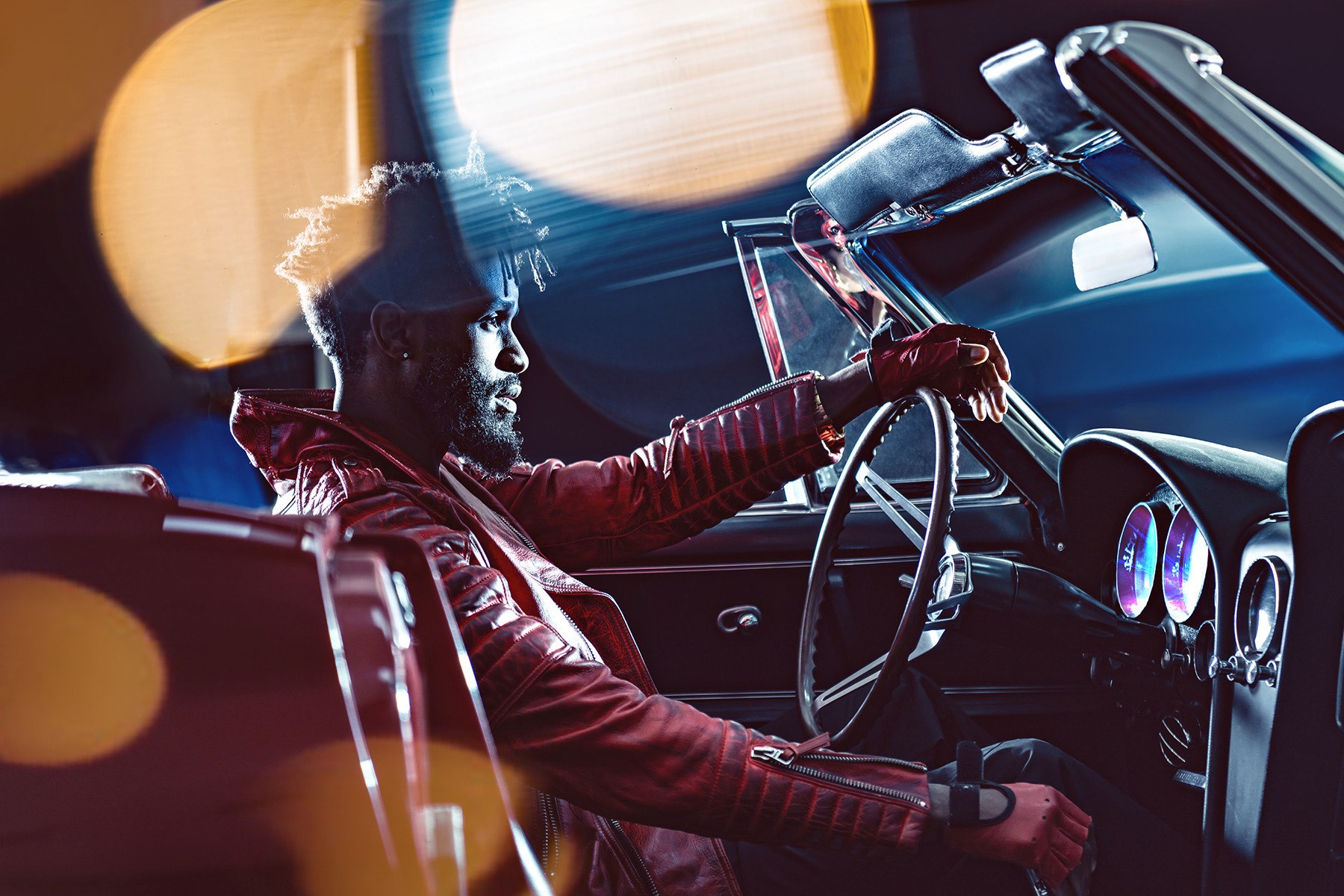 Person driving a car at night with city lights in the background, wearing a jacket and glasses, illuminated by dashboard lights.