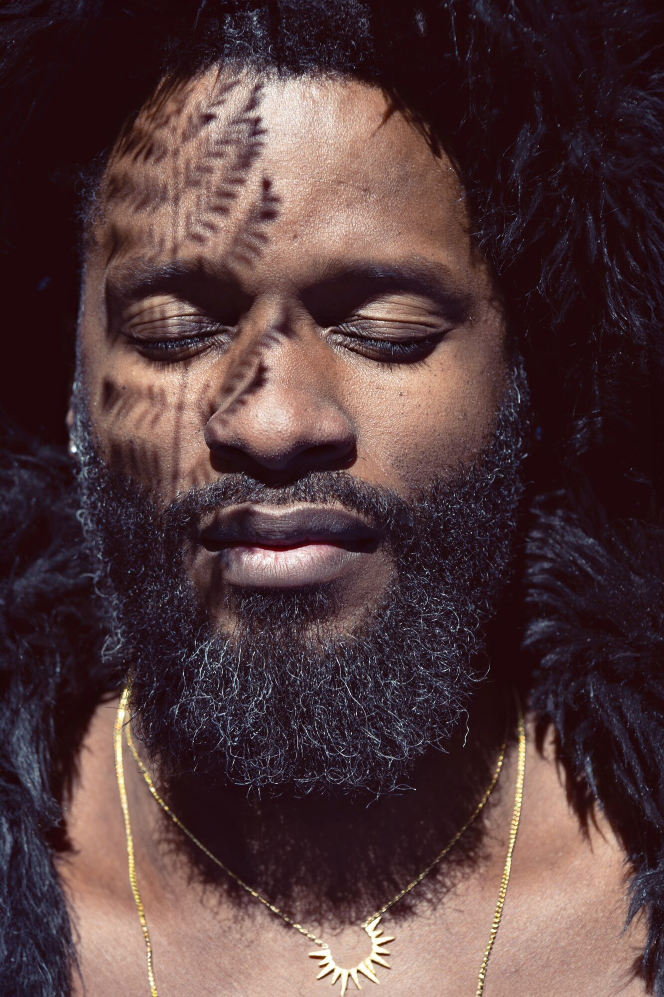 Close-up of a man with closed eyes, shadow patterns on face, beard, and necklace, with dark background.