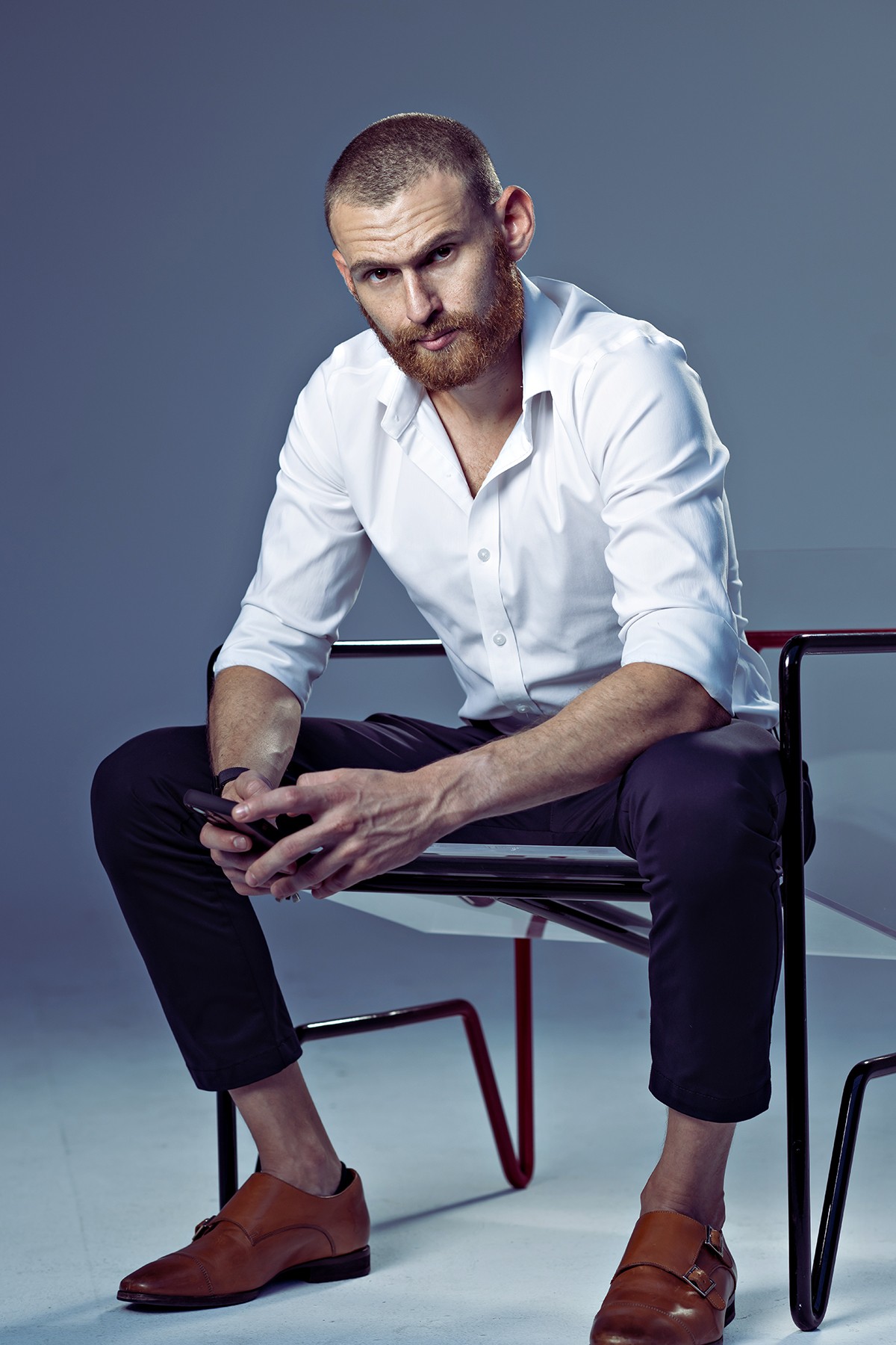 Man with a beard and short hair, wearing a white shirt and brown shoes, sitting on a chair against a plain background.