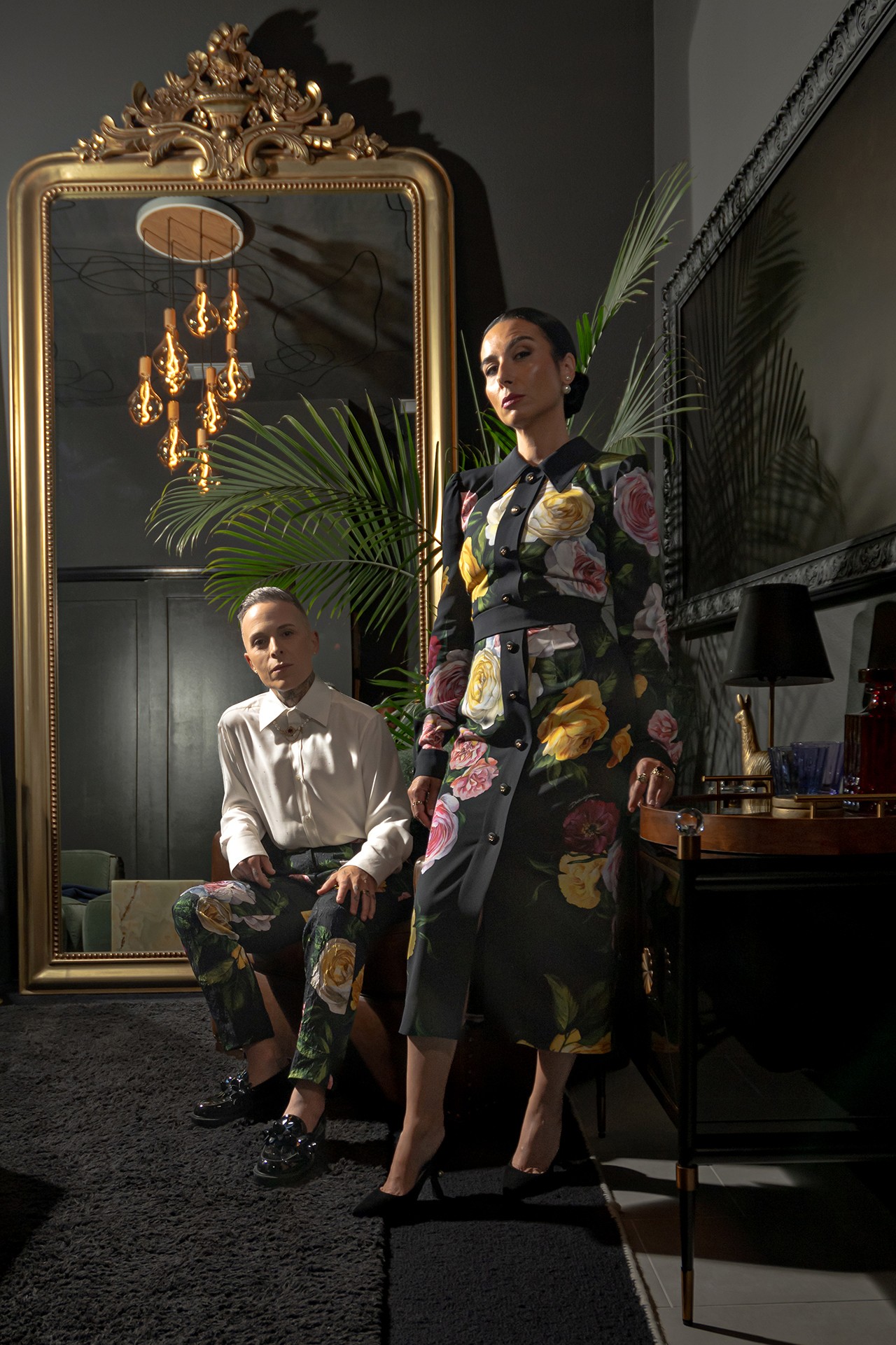 Two women pose in front of a large mirror with a gold frame, one standing and one sitting, in a dimly lit room.