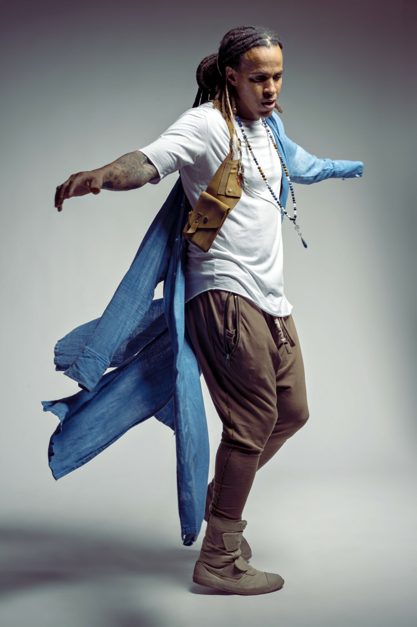 Person with dreadlocks wearing a white shirt, brown pants, and a blue jacket, standing with arms outstretched.