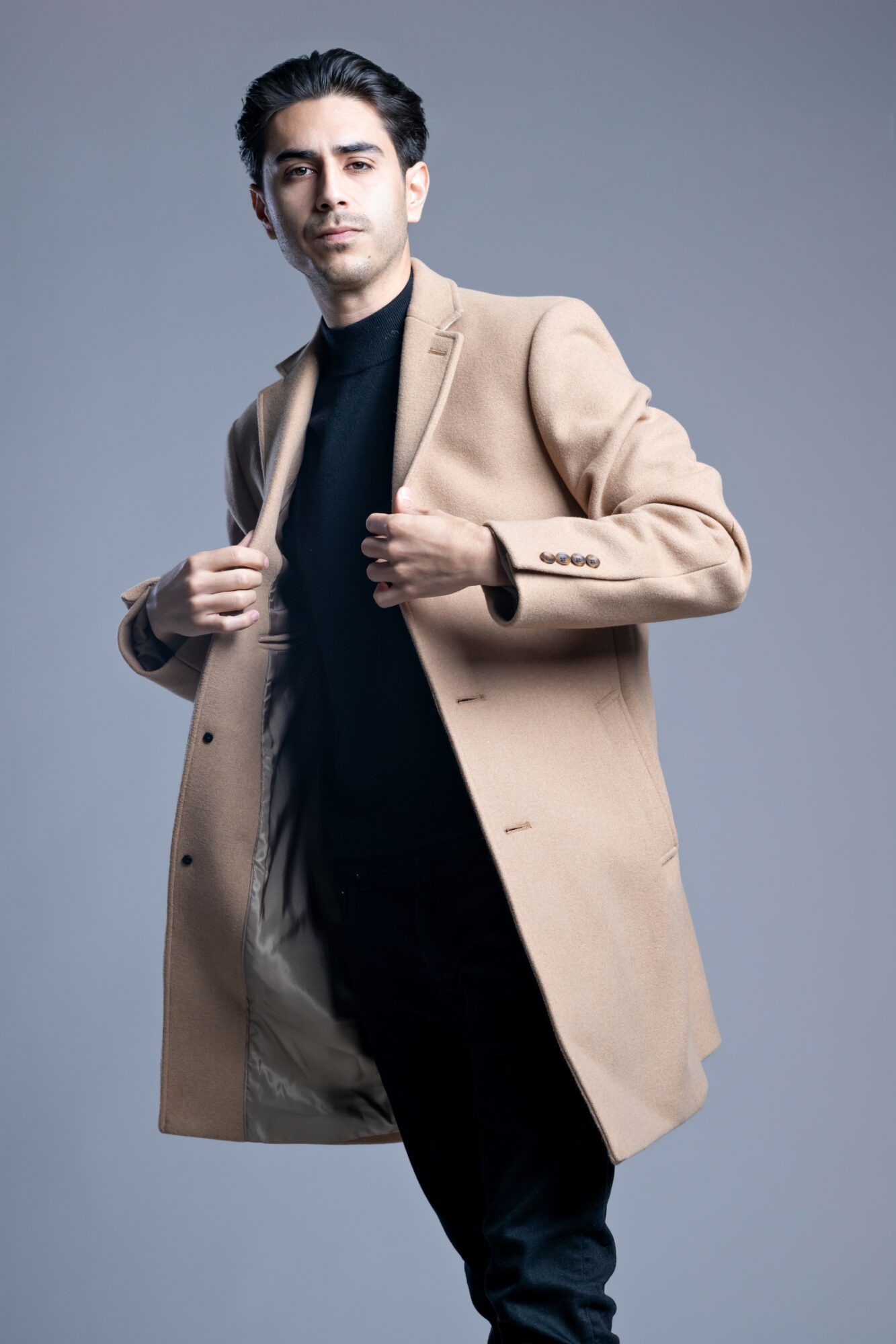 Young man in beige coat and black outfit poses confidently against gray background.