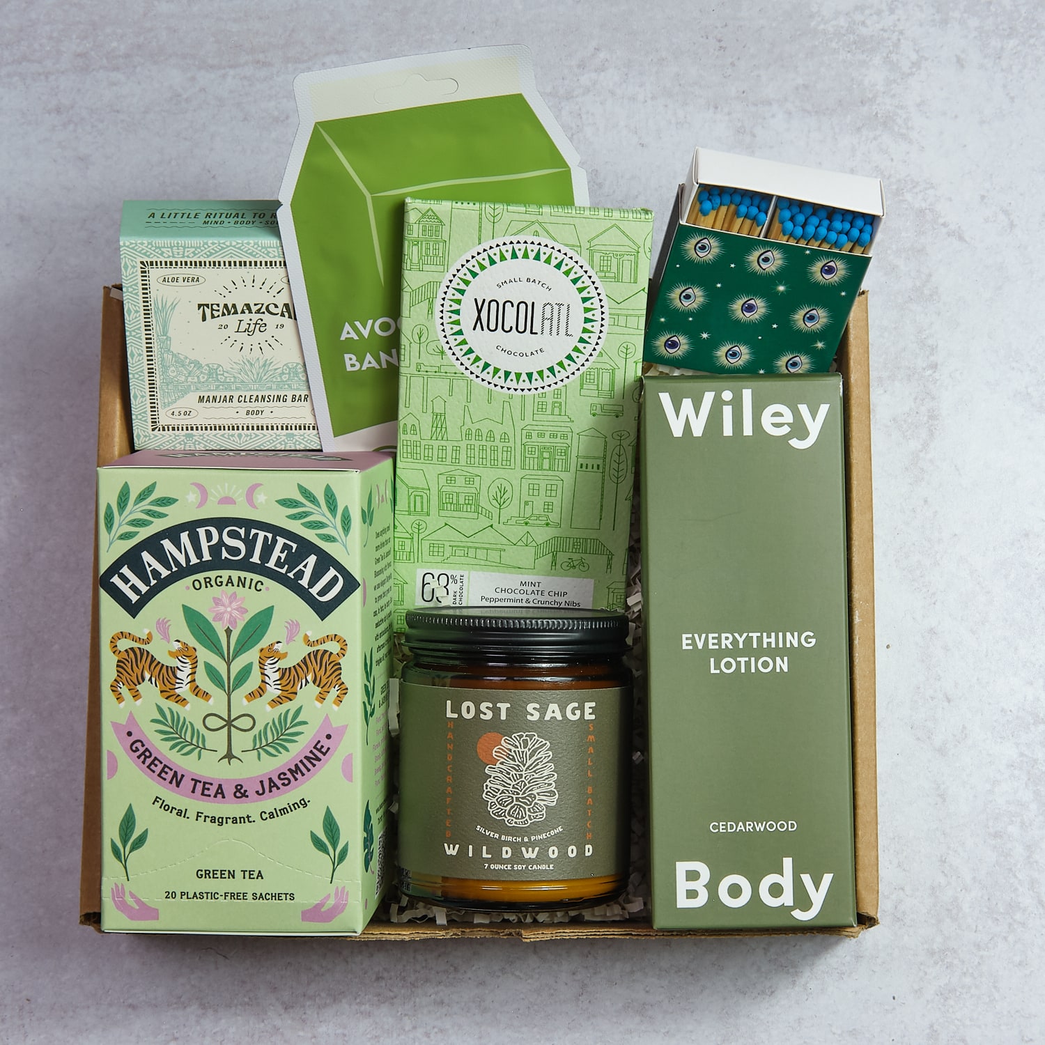 Open box containing green tea, herbal products, a candle, and lotion boxes on a gray surface.