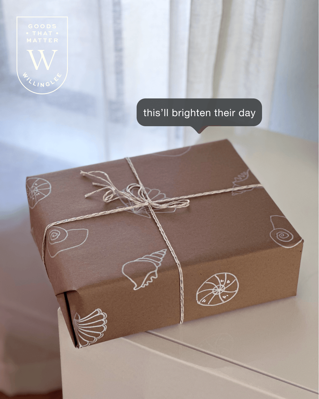 A wrapped gift box with white string and shell illustrations on brown paper, placed on a white surface near a window.