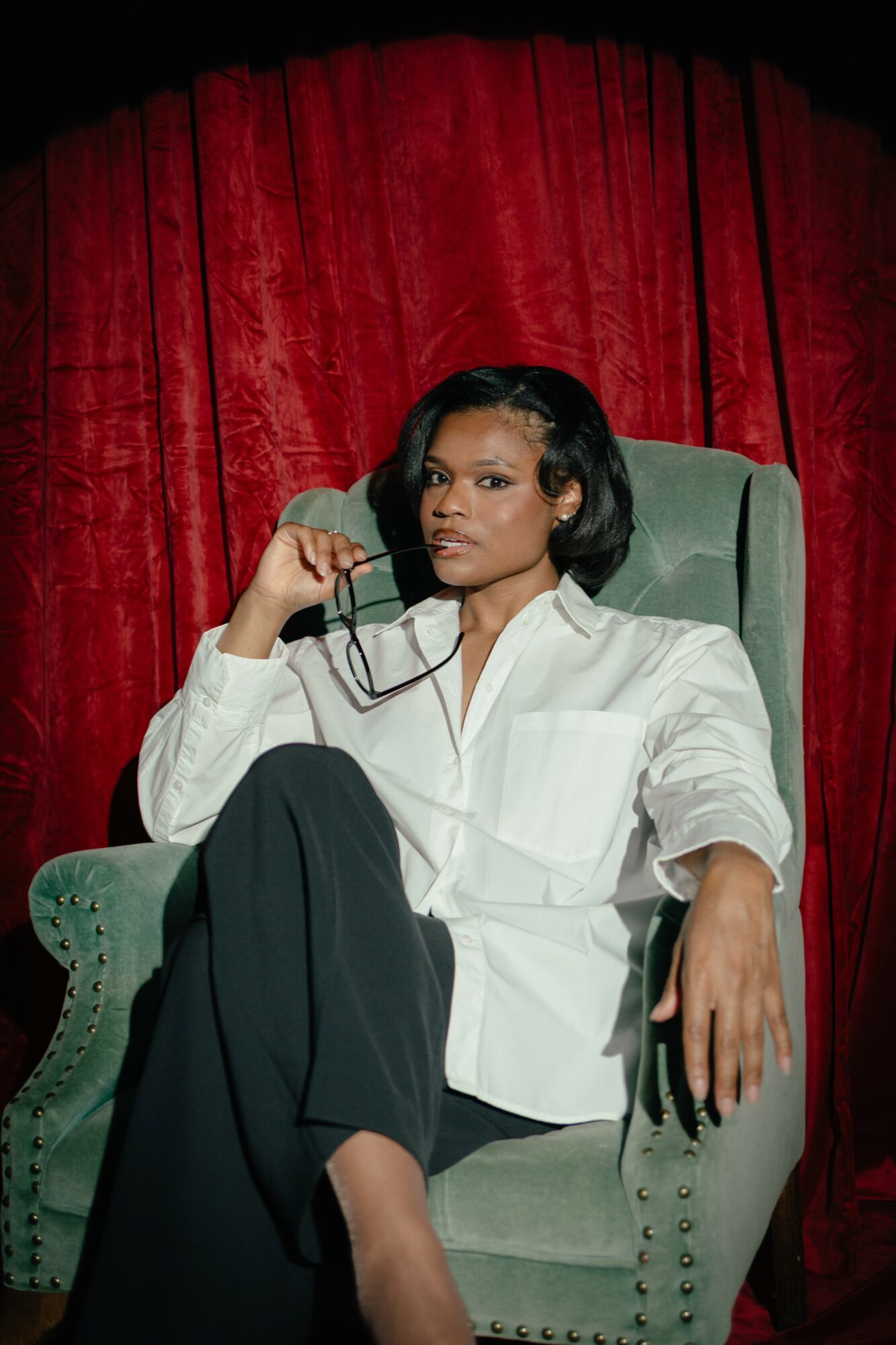 Woman sitting in a green armchair with a red curtain background, wearing a white shirt and black pants, holding glasses.