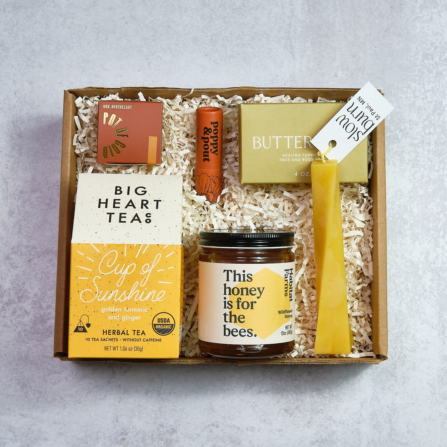 Gift box with tea, honey, soap, and a yellow candle, arranged on shredded paper. Text on items includes 'Big Heart Teas' and 'This honey is for the bees.'